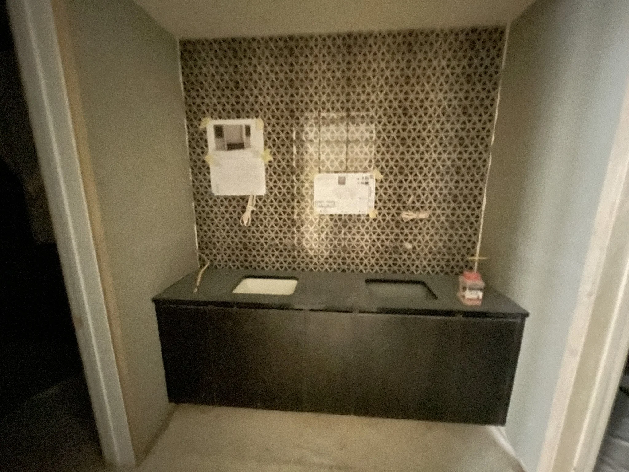 Empty kitchenette with black cabinets, a small sink, and a countertop, with a patterned backsplash and wall-mounted control panel and paper notices.