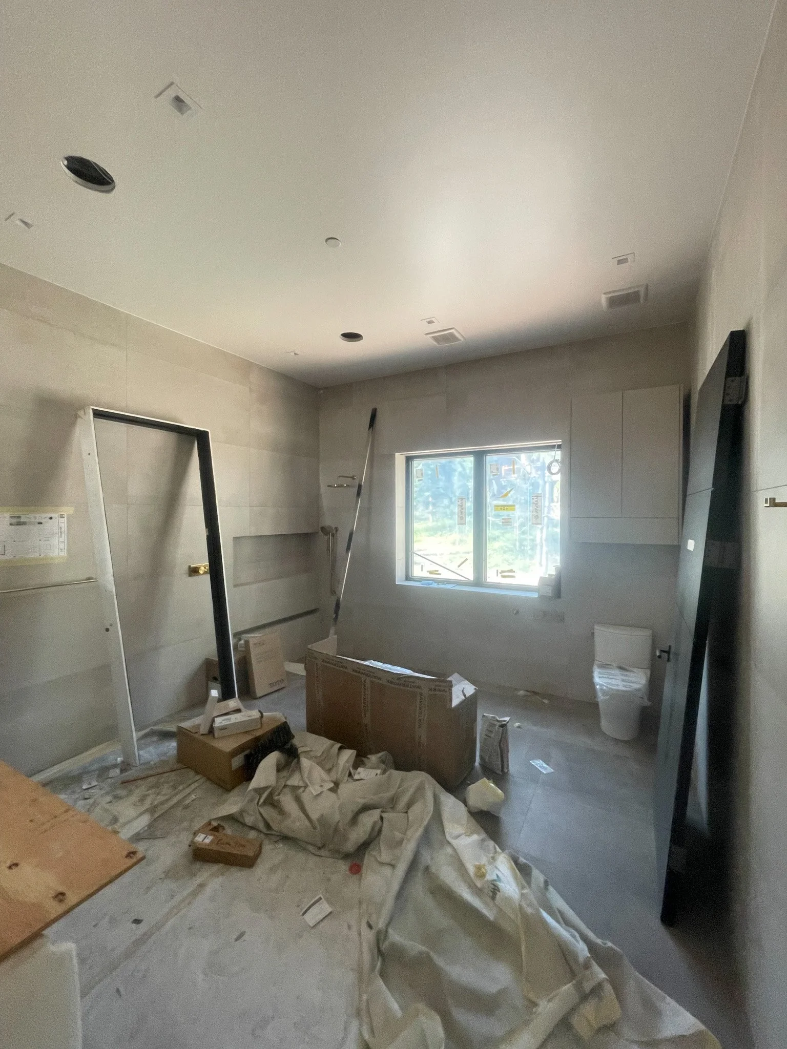 A room under construction with tools, boxes, and building materials, a wall-mounted toilet, and a window letting in natural light.