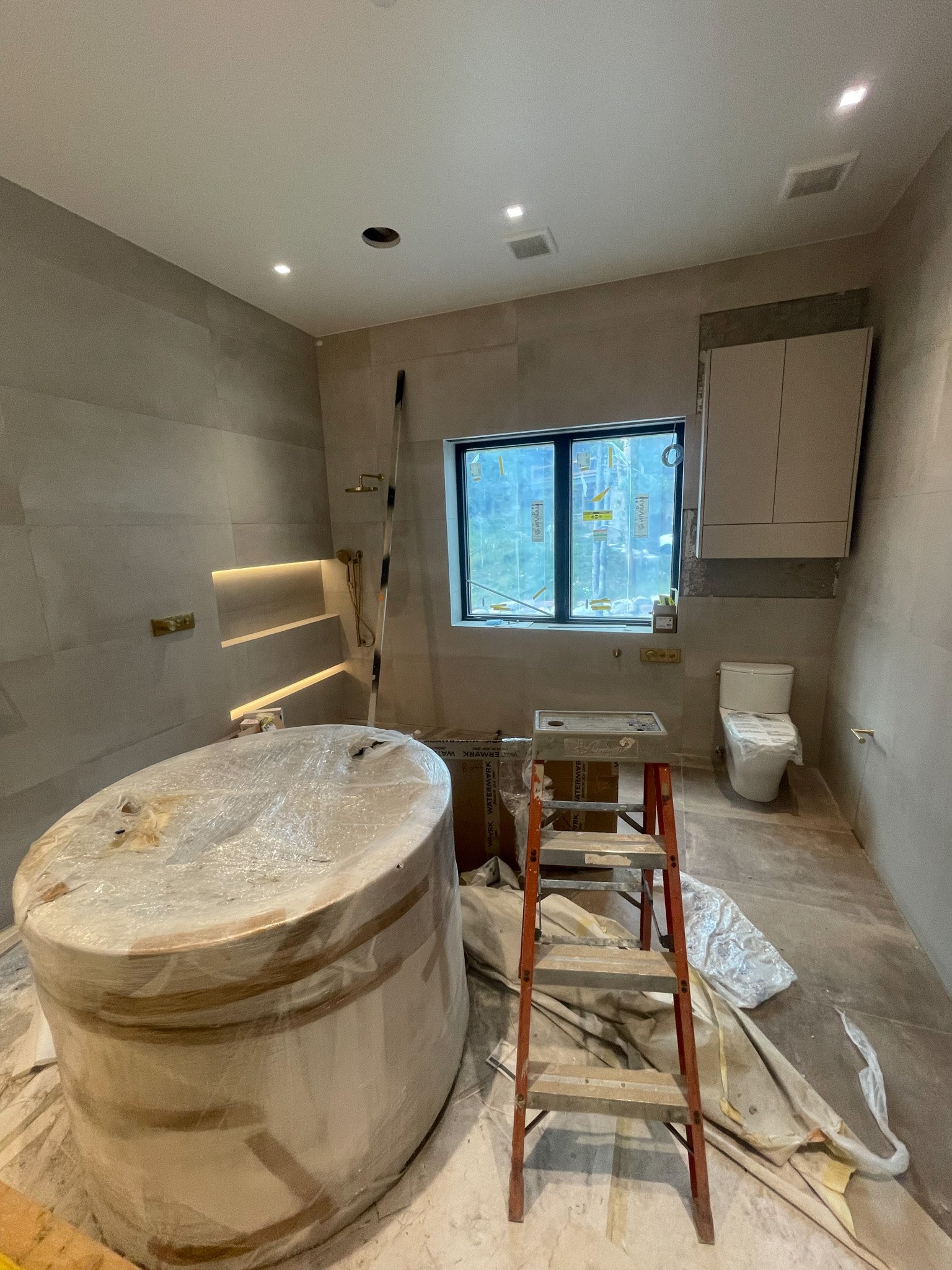 Bathroom under construction with a bathtub covered in plastic, a step ladder, a wall-mounted cabinet, a toilet, a window, and construction materials and tools scattered on the floor.