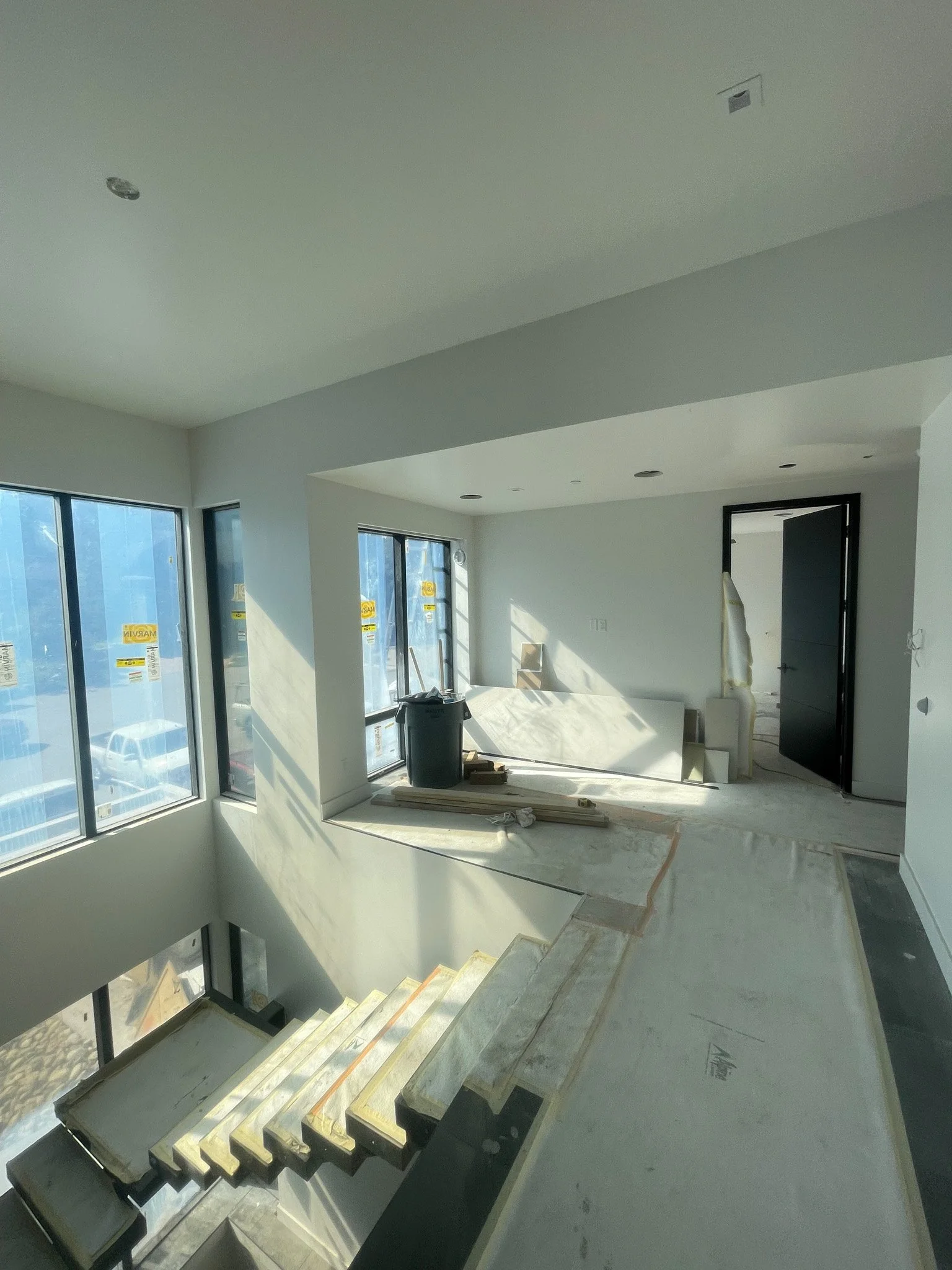 Interior view of a house under construction with a staircase, large windows, and unfinished floors. Construction materials and tools are present in the space.