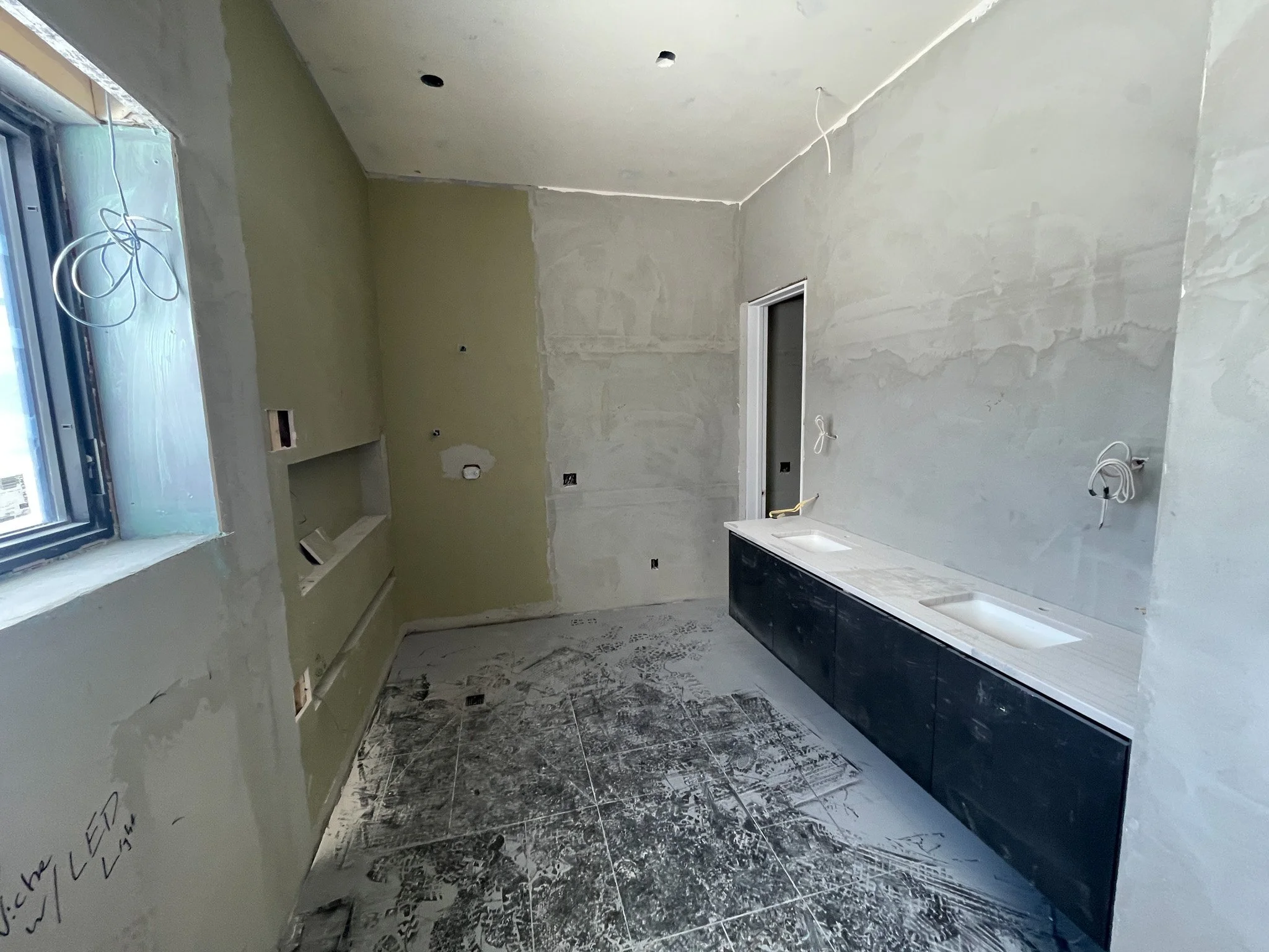An unfinished bathroom under construction with blue-tiled window, green wall, gray unpainted walls, and a long black vanity with built-in sinks and marble countertop, with electrical wires hanging from the ceiling and walls.