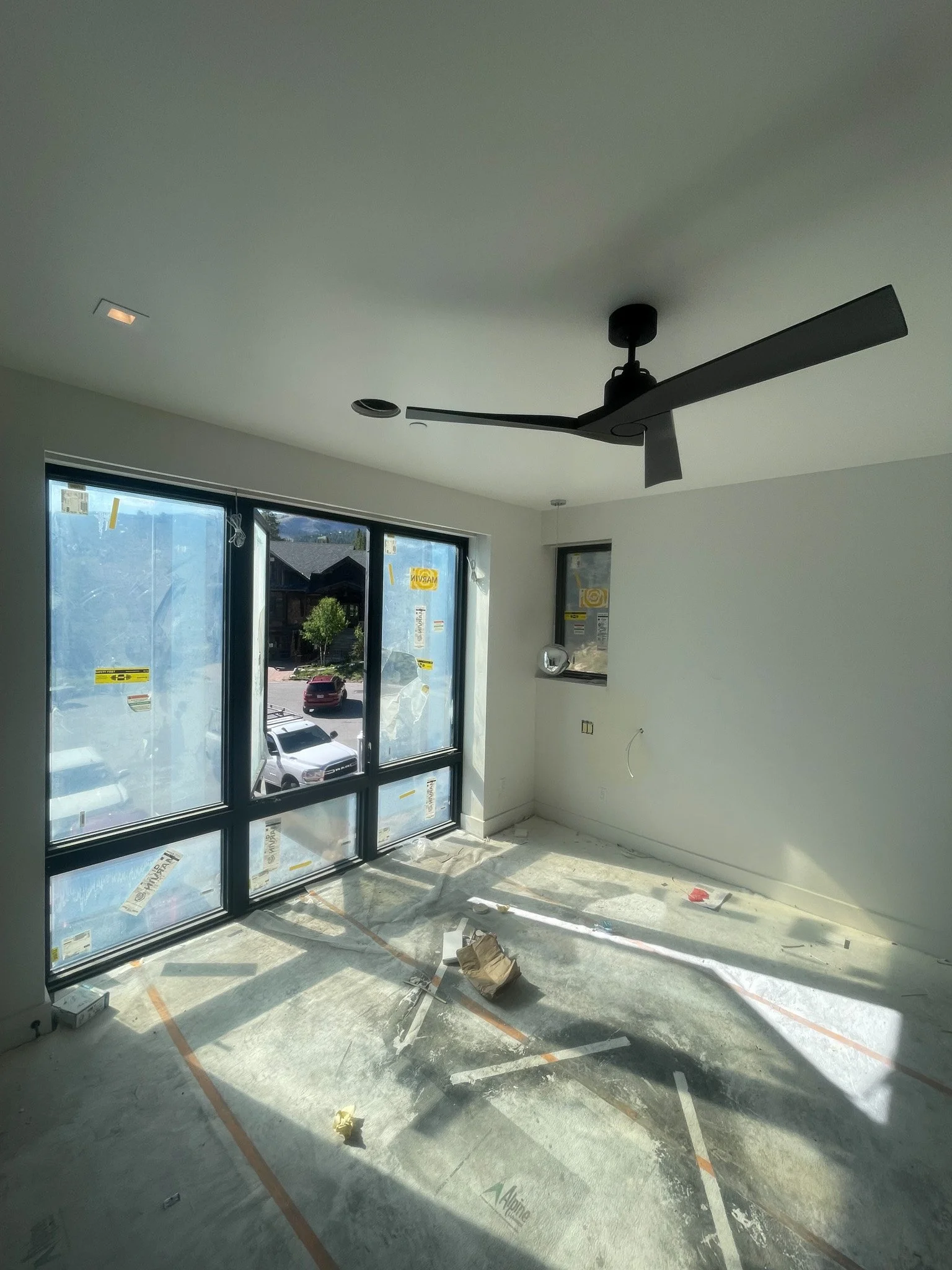 Empty room with large window showing outside parking lot, construction debris on floor, ceiling fan, and small window in corner.