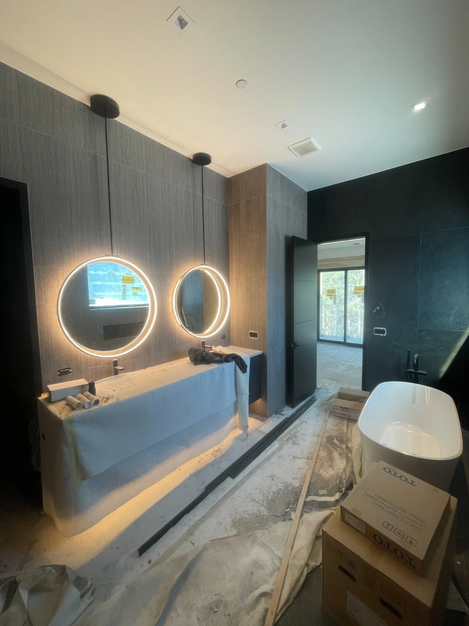 Bathroom under construction with two illuminated round mirrors above a counter, a door open to a balcony, and construction materials on the floor.
