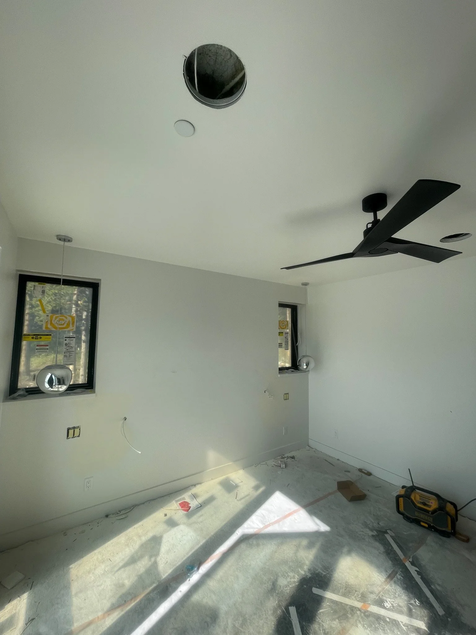 Empty room under construction with white walls, two windows, a black ceiling fan, and unfinished flooring.