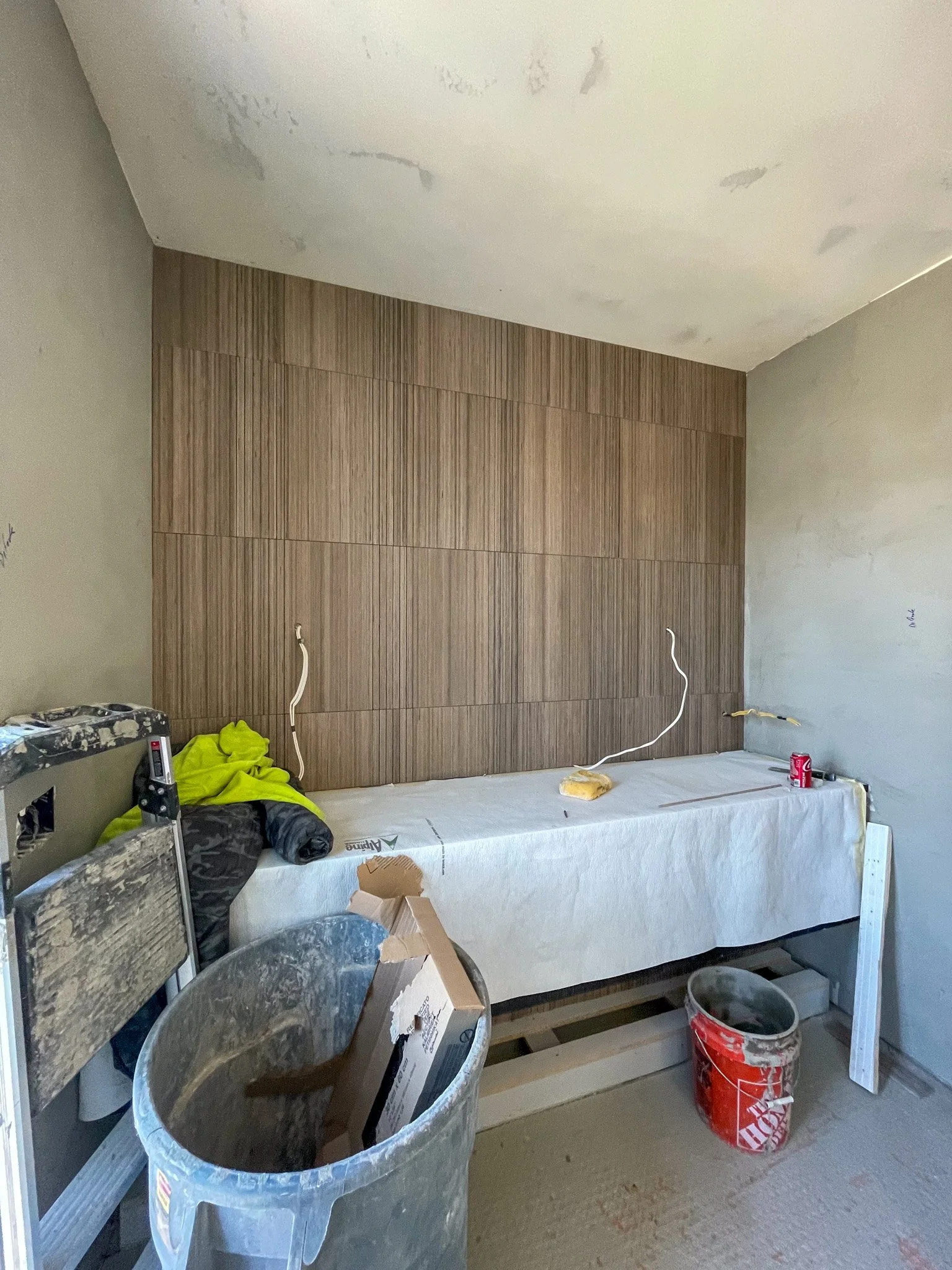 Room under construction with a wood panel wall, construction tools, a workbench covered with a white sheet, a trash can, and construction materials.