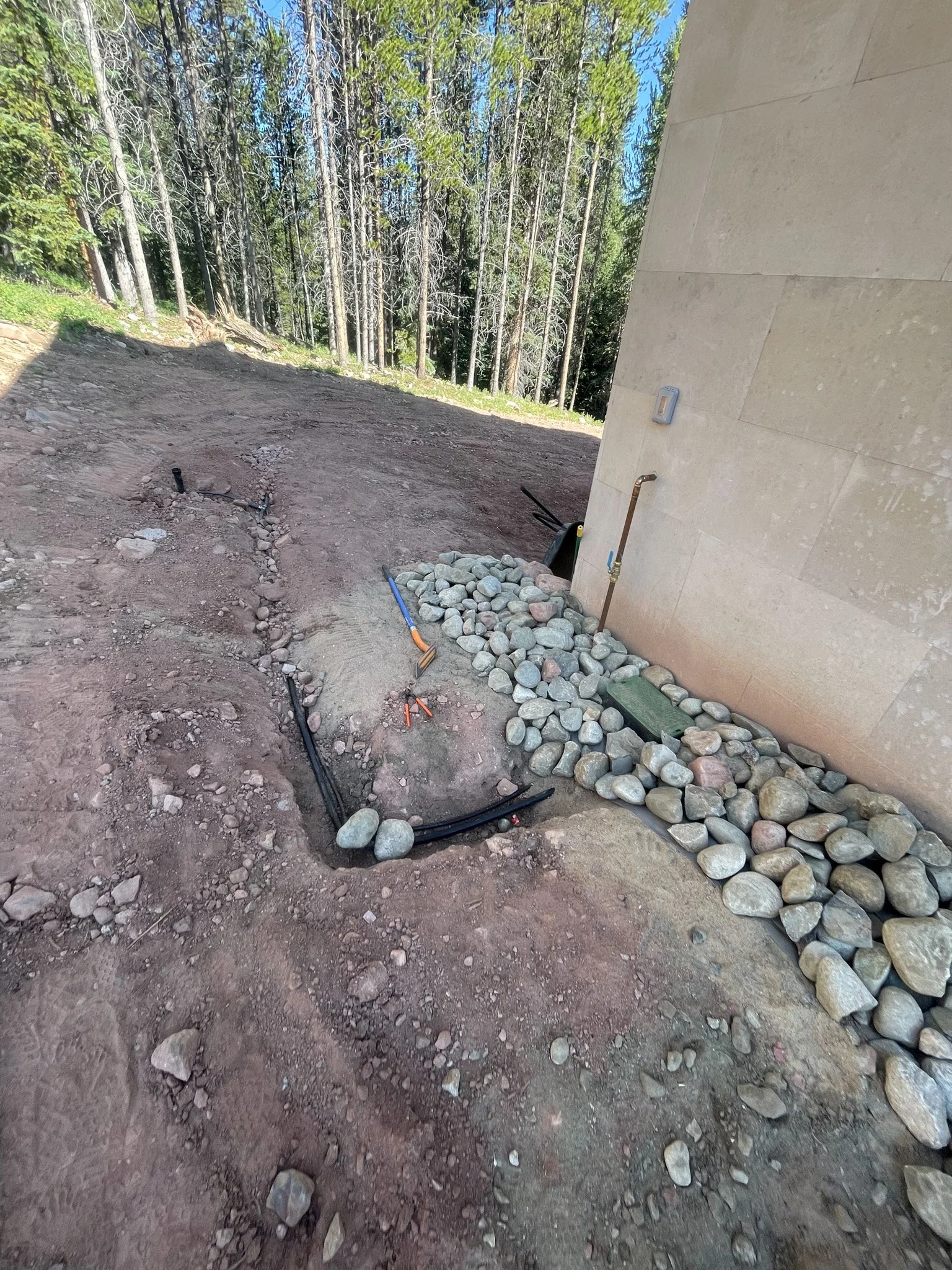 Construction site area near the corner of a building with visible pipes, rocks, and exposed soil, with trees in the background.