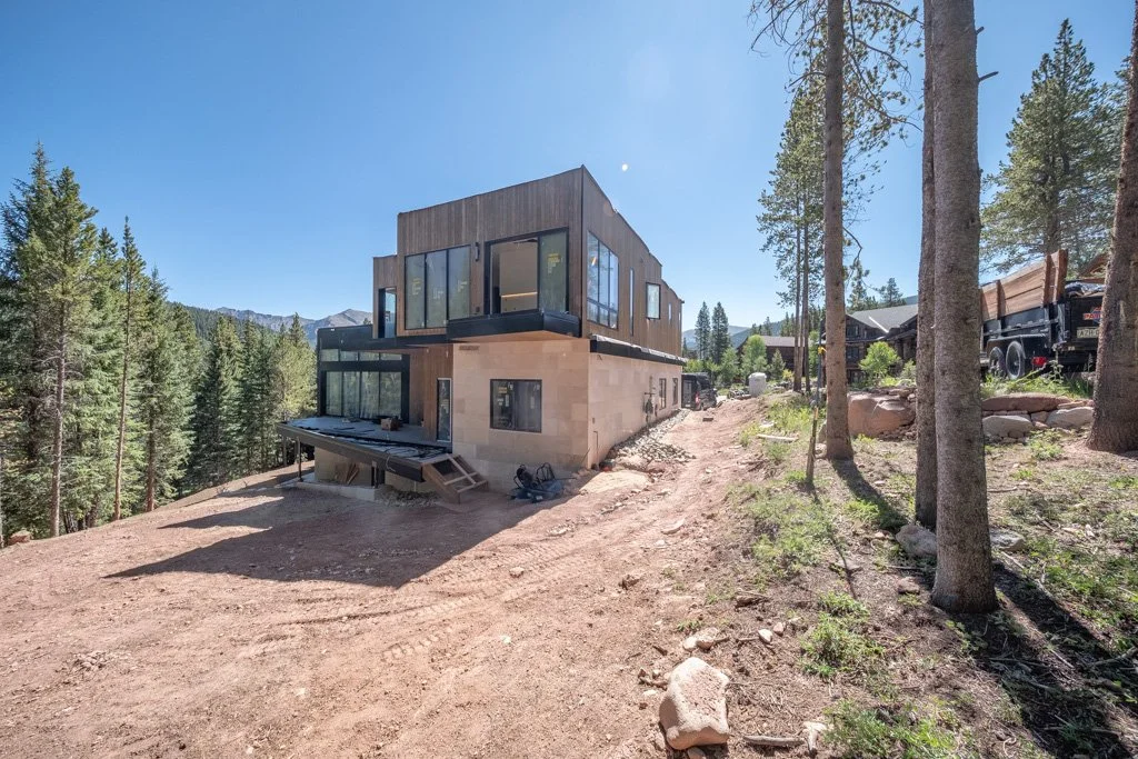 Modern house under construction on a dirt lot in a wooded area with mountains in the background.
