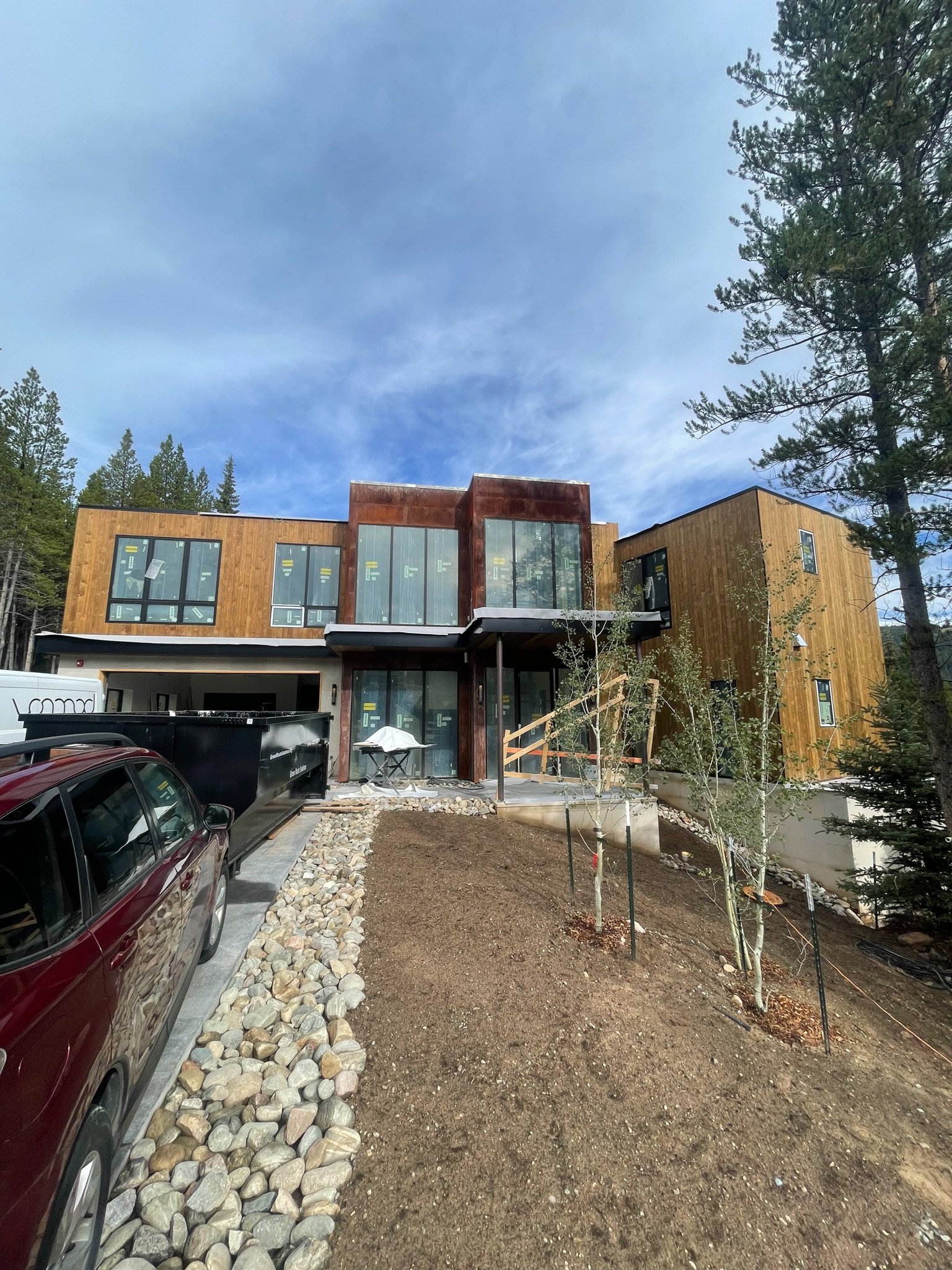 Modern two-story house under construction with wooden and rust-colored exterior, large windows, and a driveway with cars and rocks, set against trees and a partly cloudy sky.