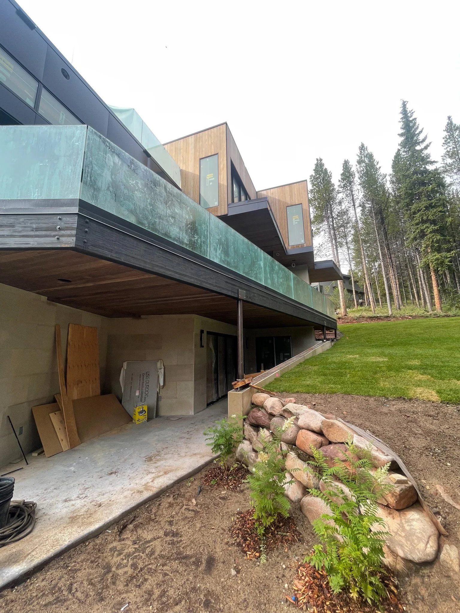Modern house with large glass balcony, wooden and black exterior, surrounded by trees and green lawn, under construction.
