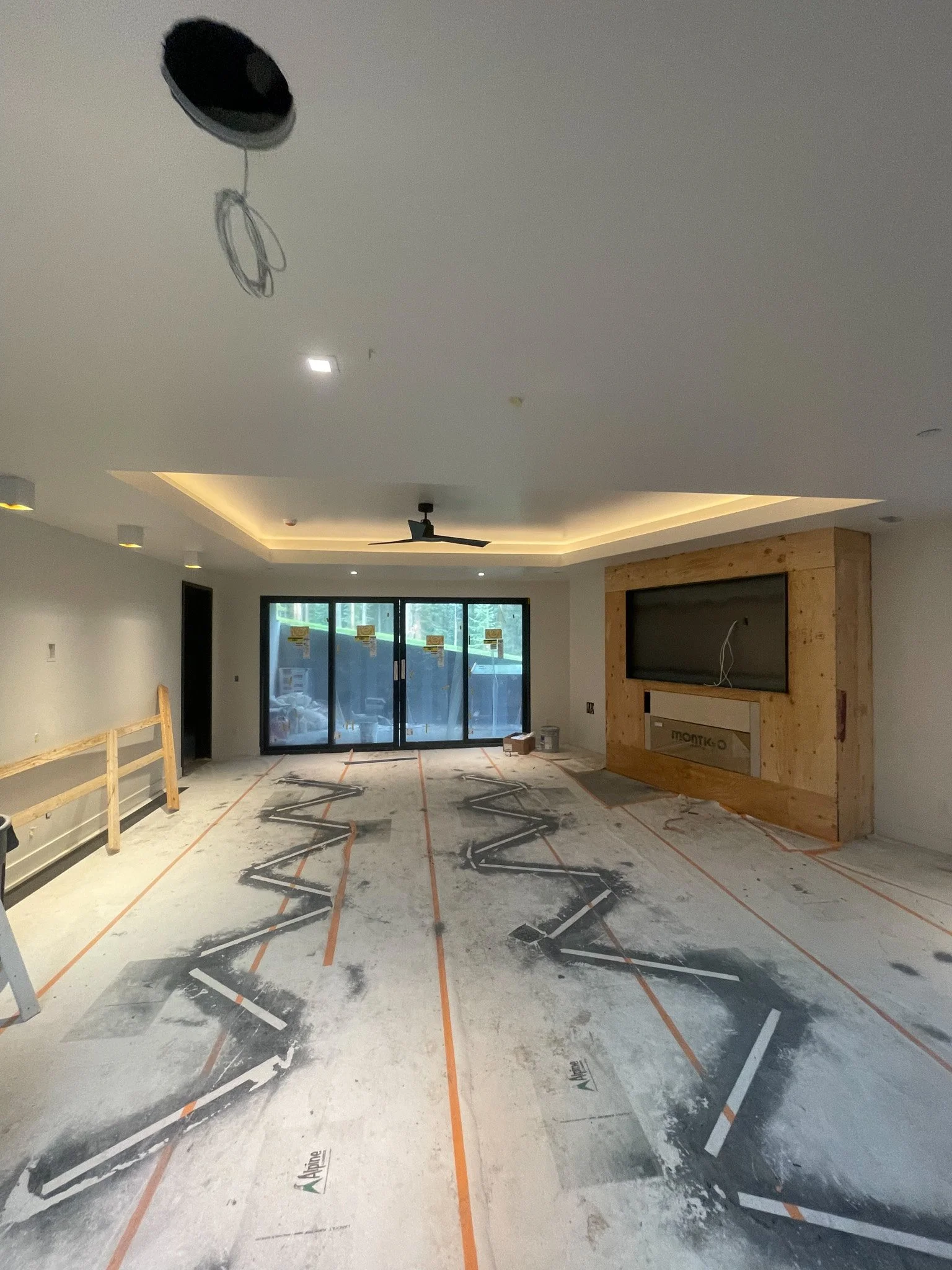 An unfinished living room during construction with a large sliding glass door, a wooden frame around a wall area, a ceiling fan, and marked flooring.