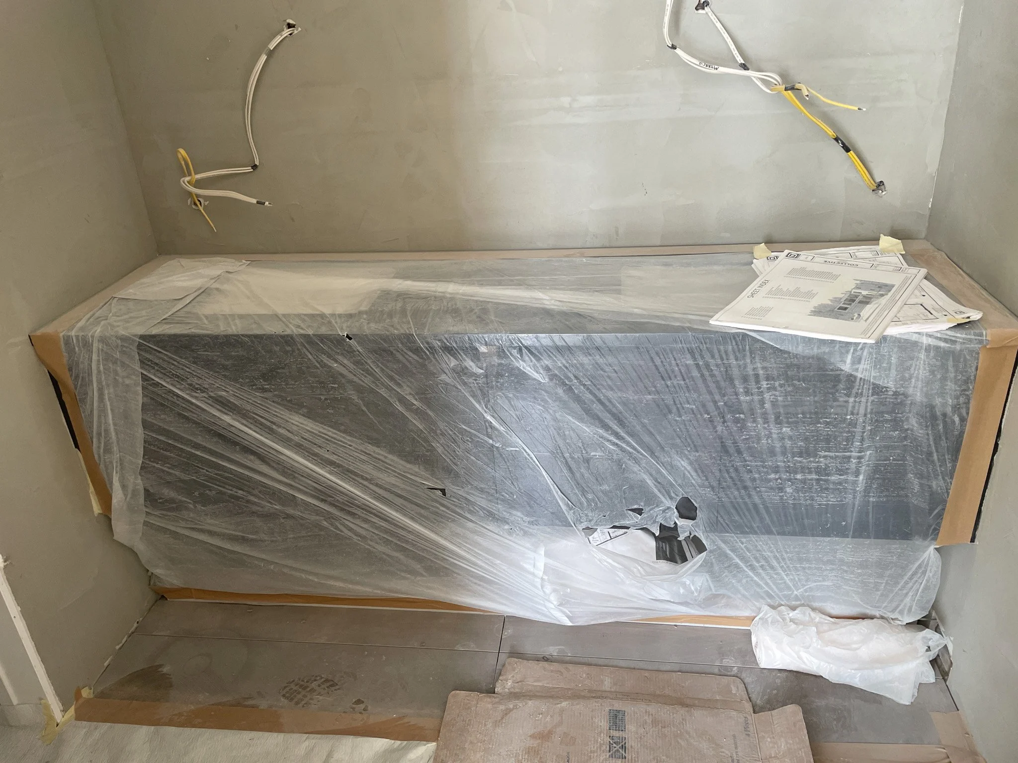 A partially covered black appliance, possibly a refrigerator, wrapped in protective plastic and tape, in a construction or renovation area. Exposed electrical wiring above and a stack of papers on top.