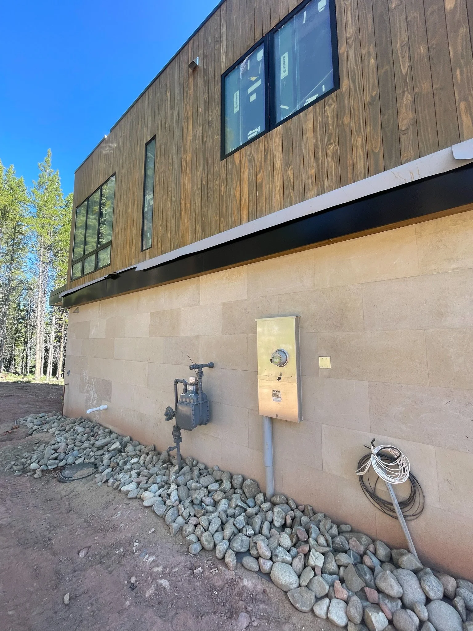 Exterior view of a modern building with wooden and stone facade, featuring large windows, utility meters, and a coiled cable on the ground.