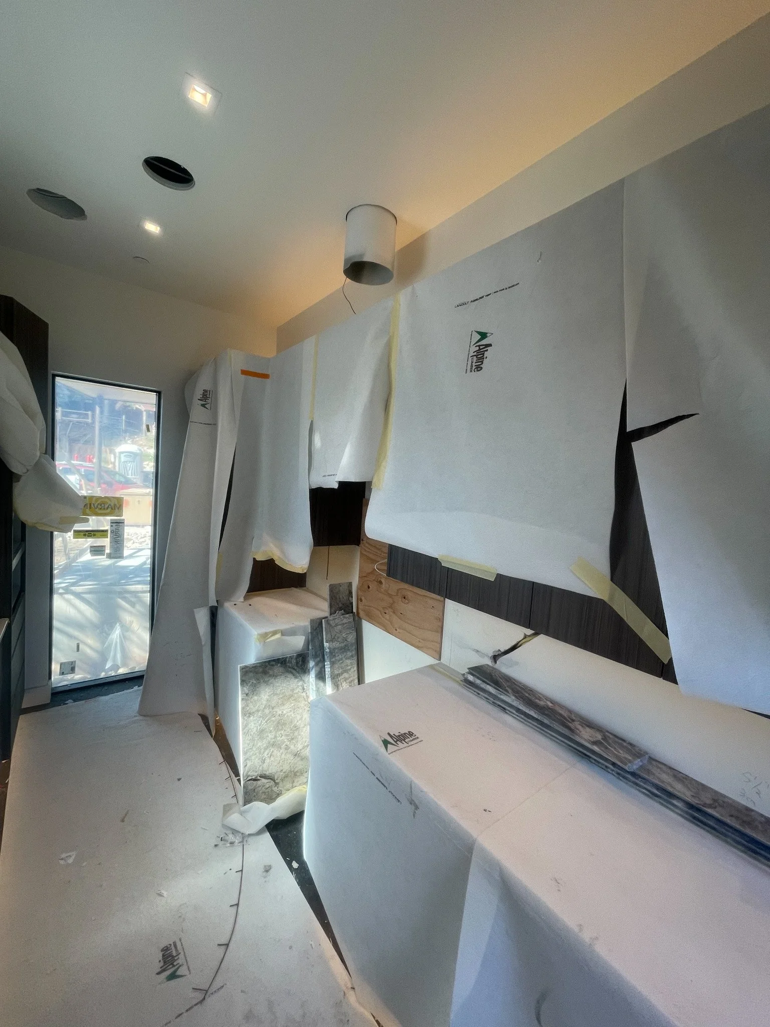 Interior of a room under construction with protective paper covering countertops and cabinets, a door with a window leading outside, and electrical fixtures in the ceiling.