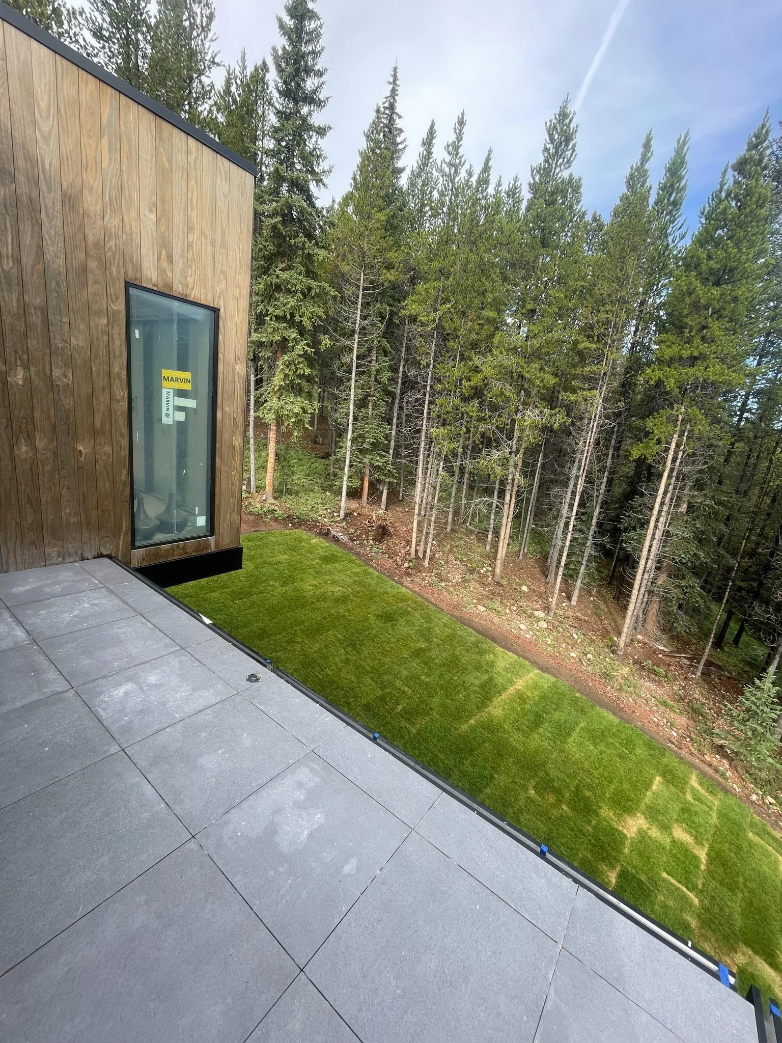 A modern building's outdoor patio with gray tile flooring, a wooden exterior wall with a large window labeled 'MARVIN', and a small patch of green grass surrounded by trees and a forested area.