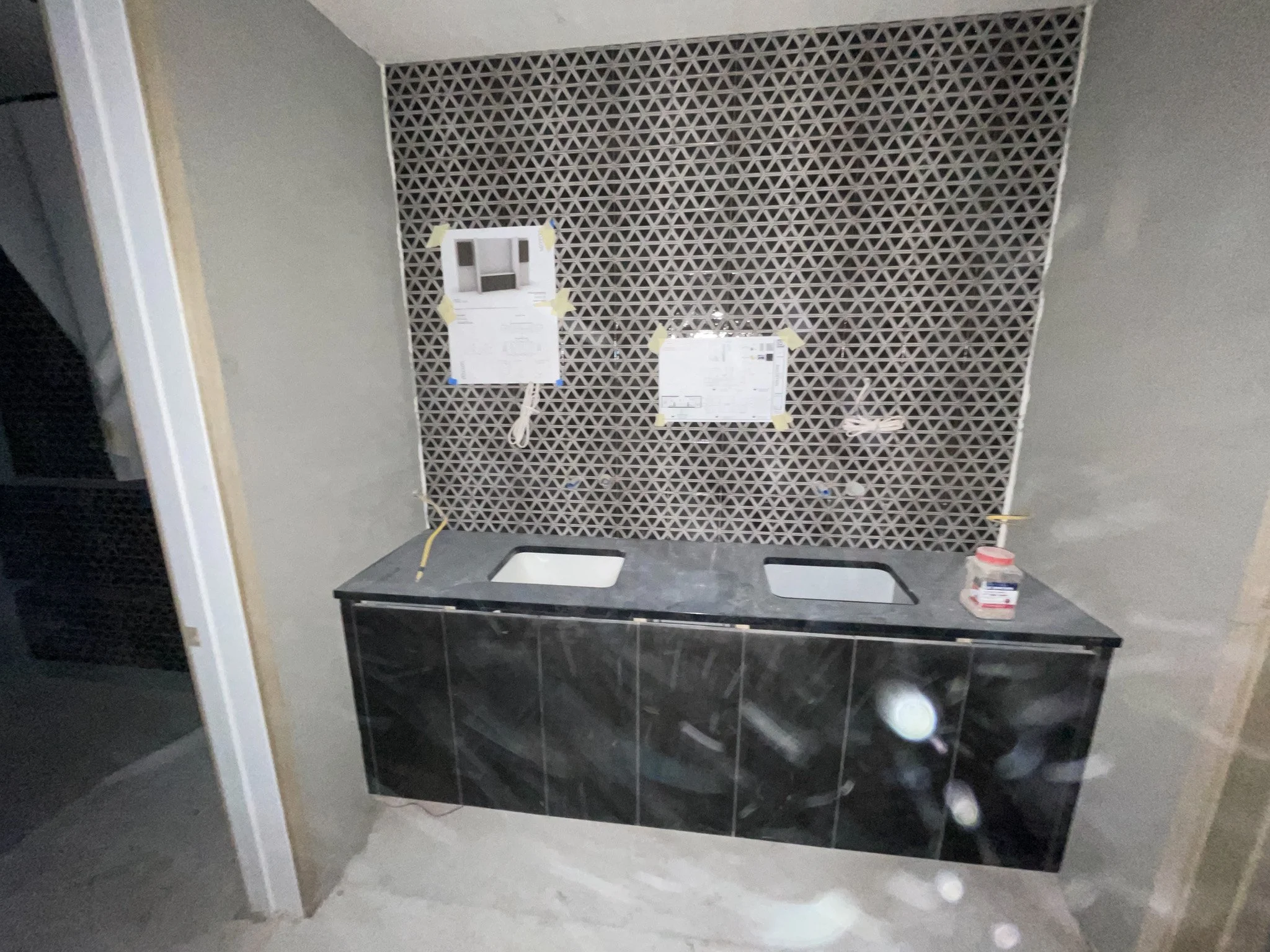 Under construction bathroom vanity with two sinks, black marble cabinet, tiled wall, construction papers taped to wall, a bottle of adhesive or sealant on the countertop.