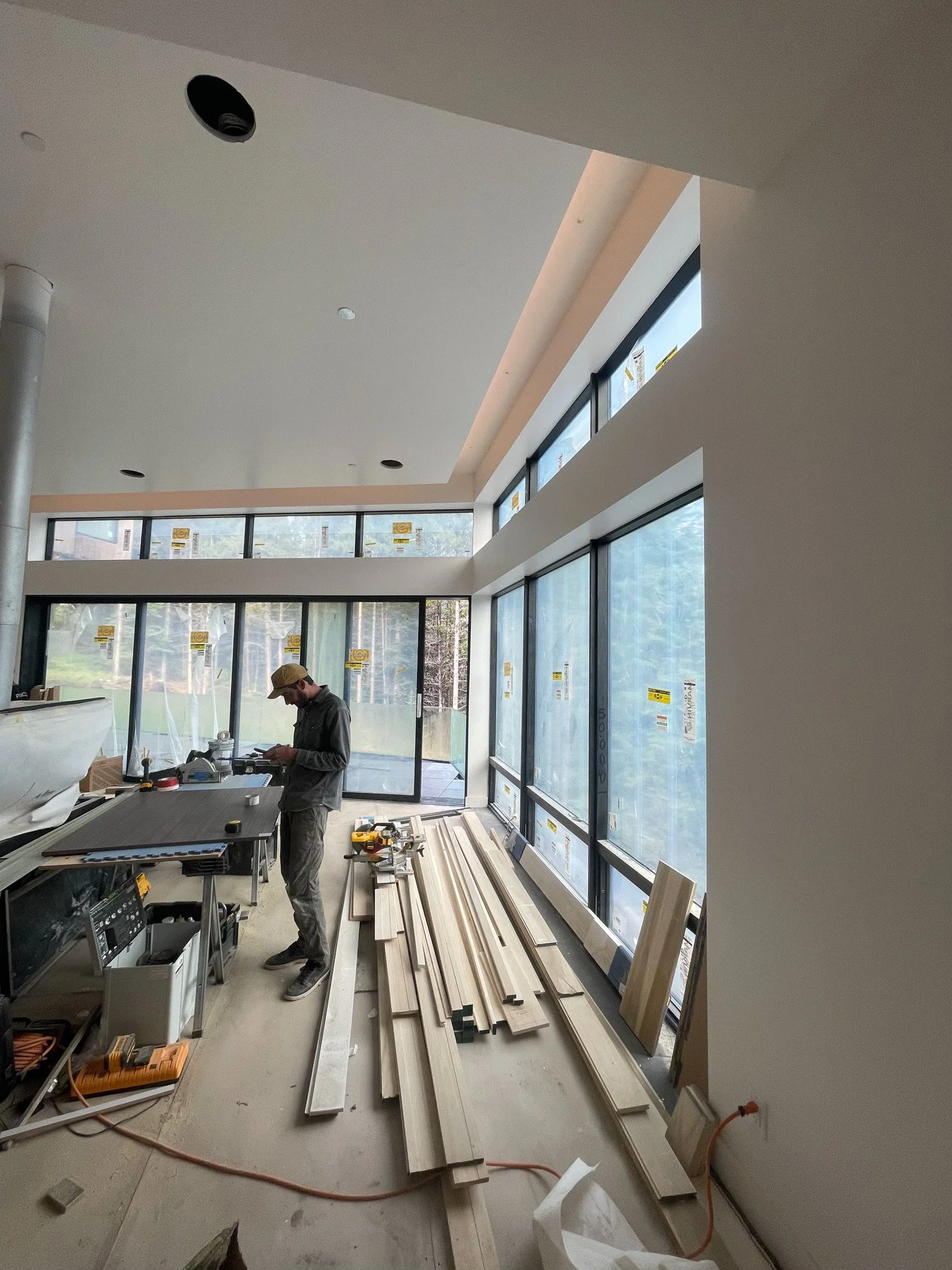 Construction worker standing in a room with large windows, working with wood and tools, with various lengths of wood and construction materials scattered on the floor.