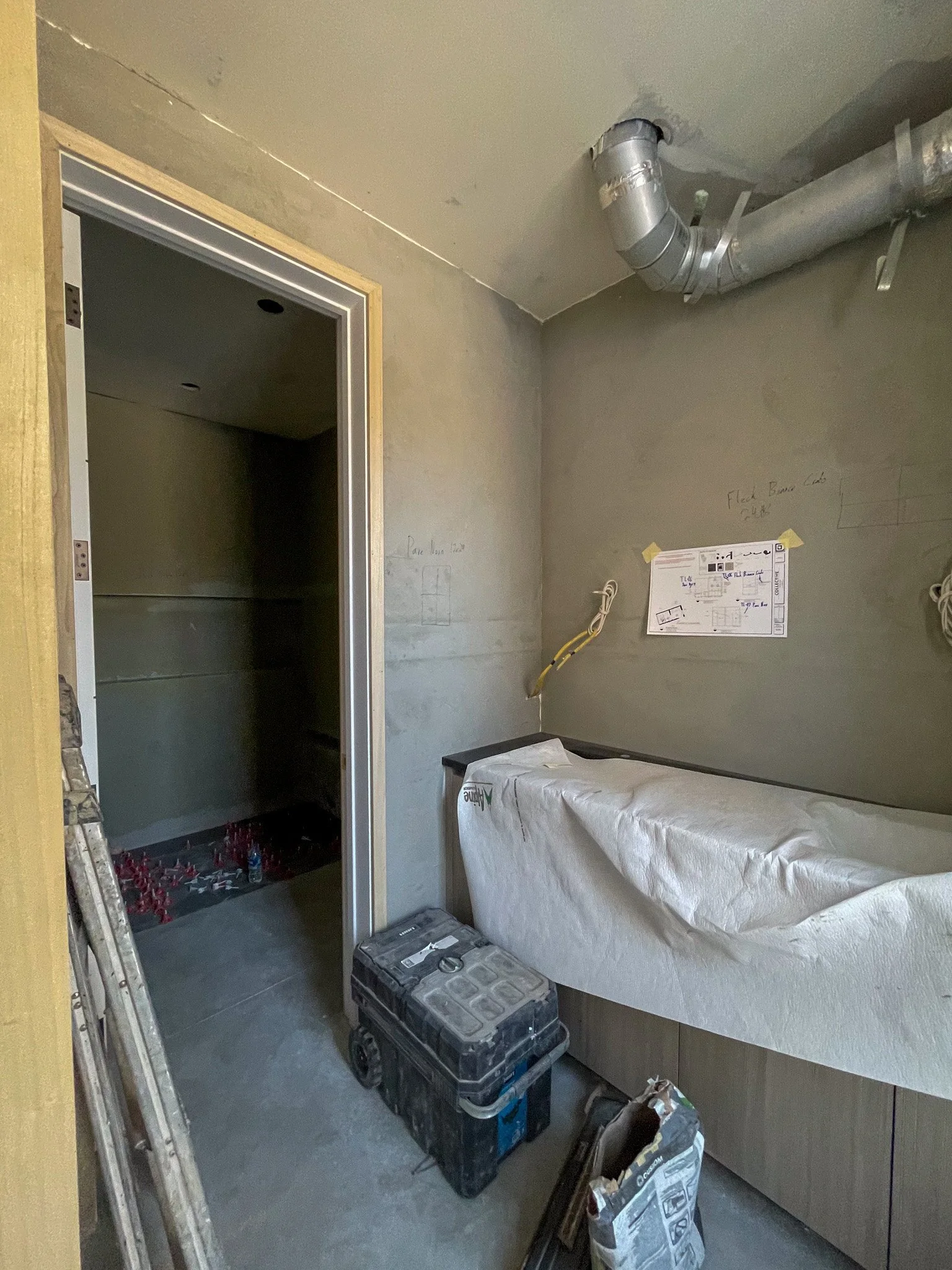 Interior of a room under construction with unfinished walls, a ceiling duct, a covered countertop, a toolbox on the floor, and construction materials and tools scattered around.