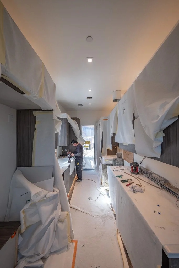Kitchen under construction with protective coverings, tools, and a worker installing or working on a countertop near the window.