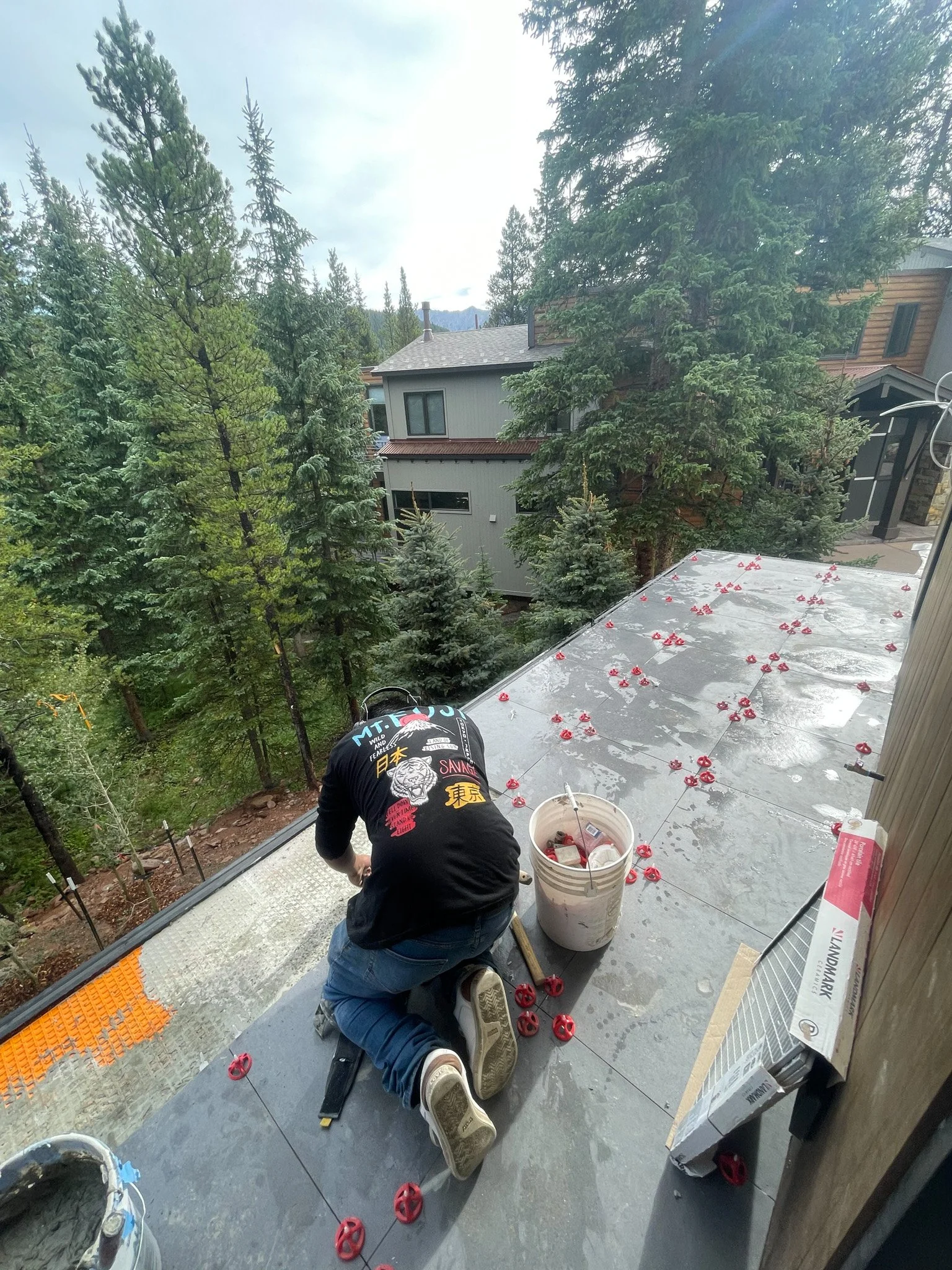 A person working on a rooftop installation or construction, kneeling down and surrounded by red plastic anchors, with a bucket and tools nearby. The rooftop has scattered red anchors, and the background shows a residential neighborhood with tall pine