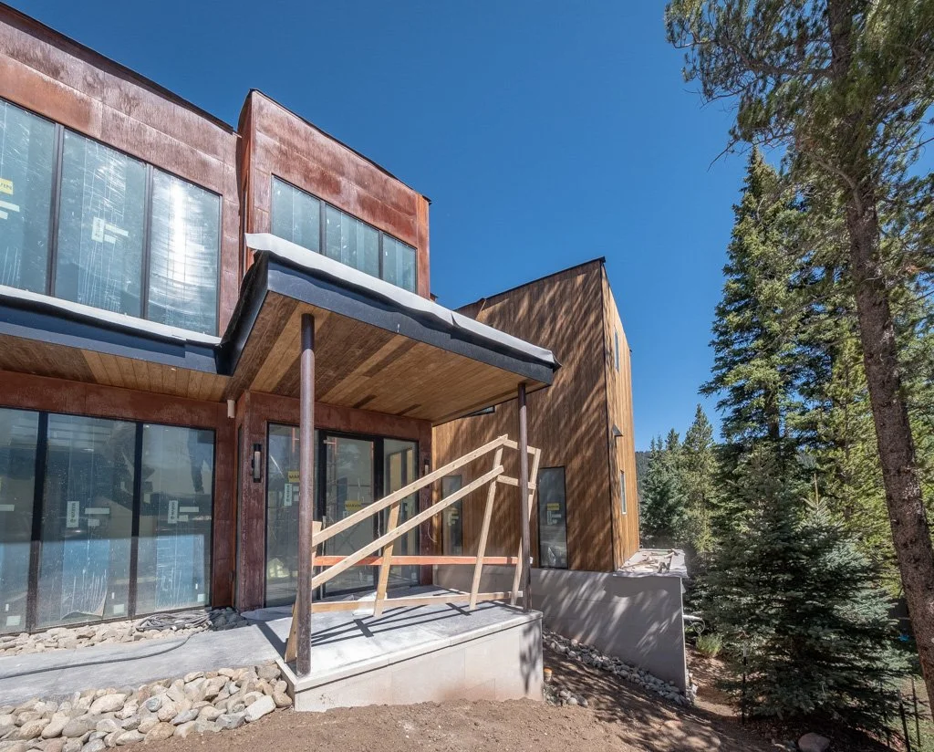 Modern house under construction with large glass windows, wooden exterior, and a small porch with a hand railing, surrounded by trees and a clear blue sky.