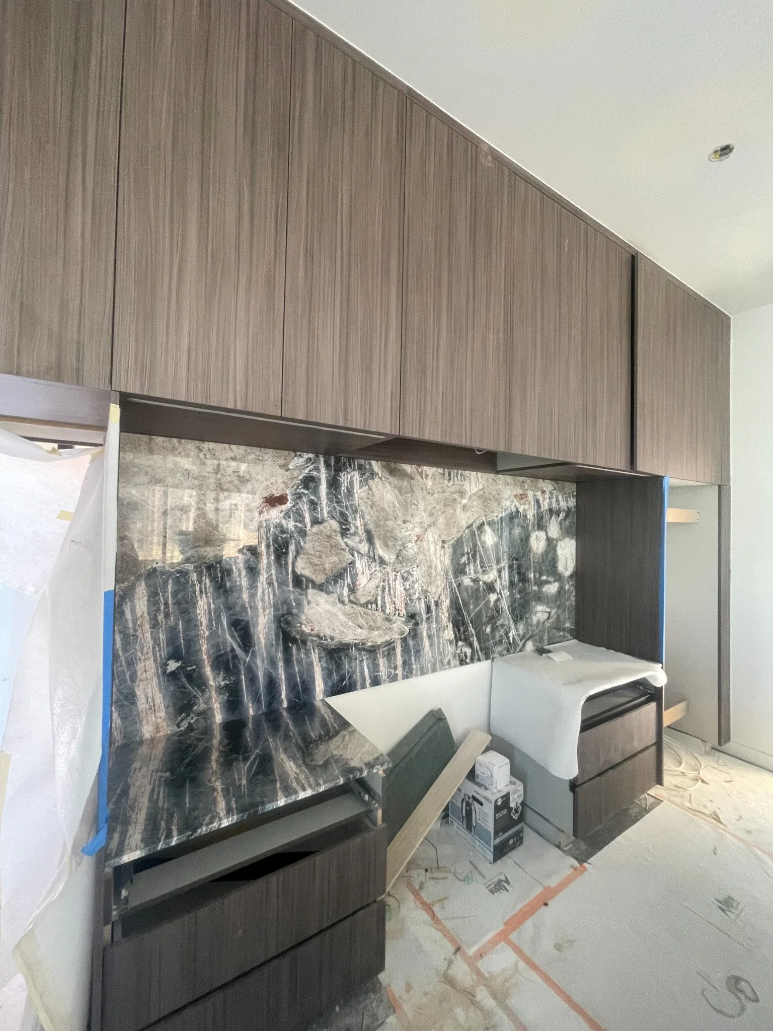 Kitchen under construction with dark wood cabinets, marble countertop with gray, white, and black veining, and protective coverings on some surfaces.