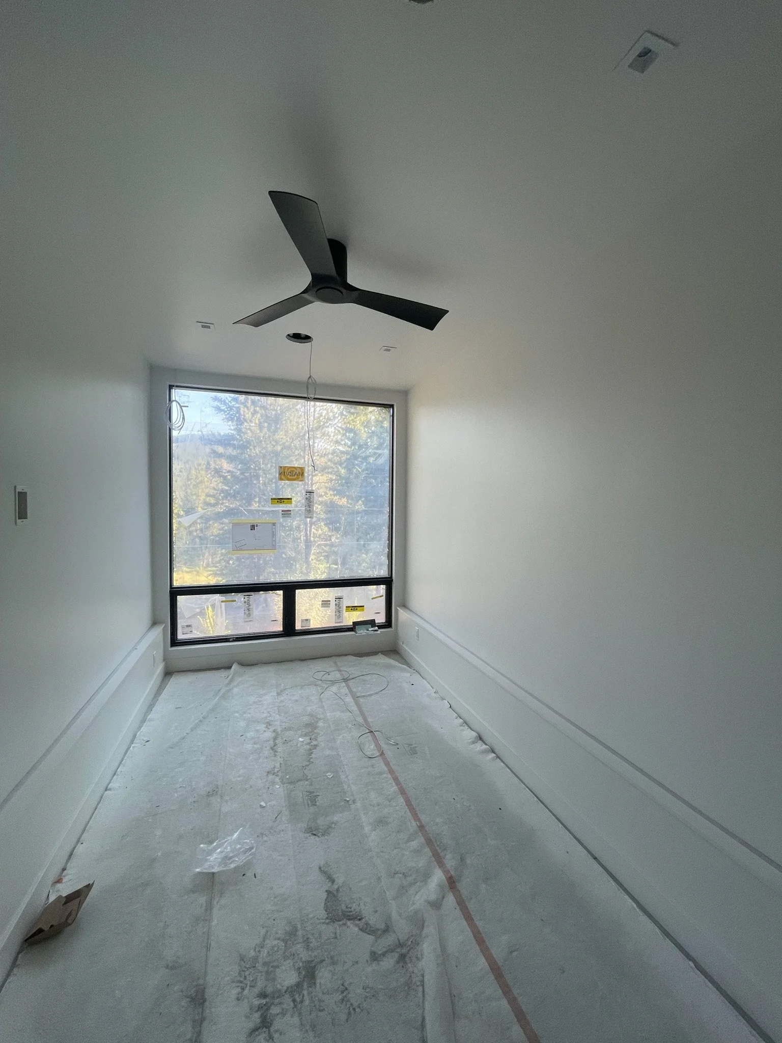 Empty room under construction with white walls, large window, ceiling fan, and unfinished floor.