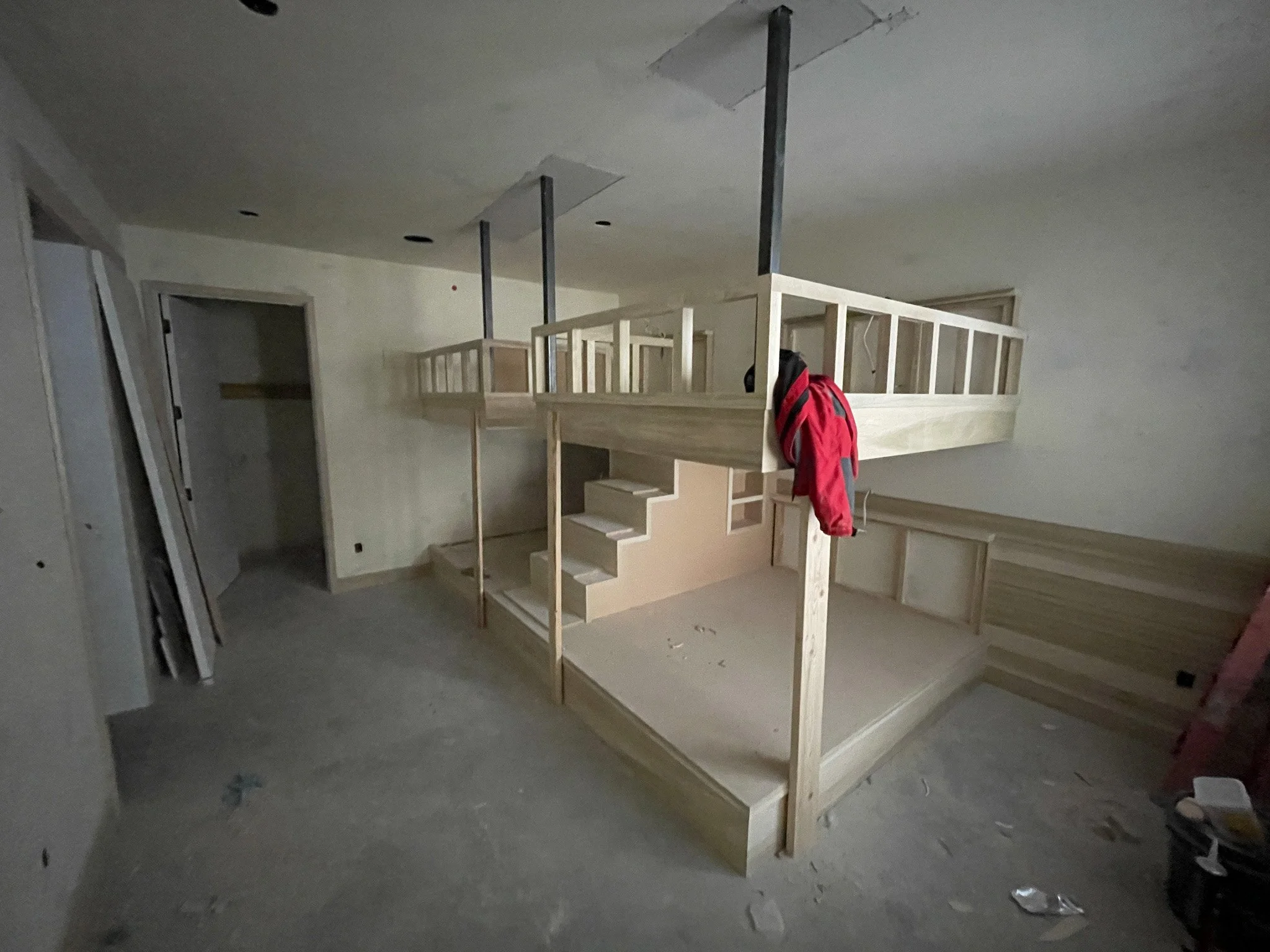 Interior view of a room under construction with a wooden loft bed frame, stairs, and various construction materials and tools scattered on the floor.