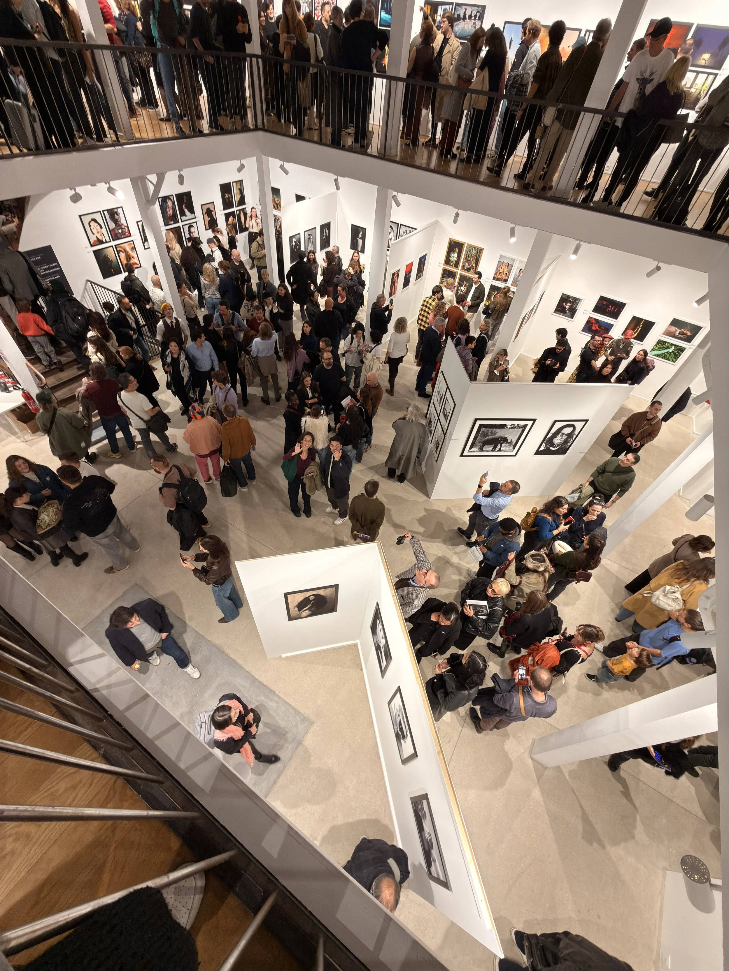 Una visita a una galleria d'arte affollata con persone che osservano fotografie e opere d'arte su pareti bianche, vista dall'alto.