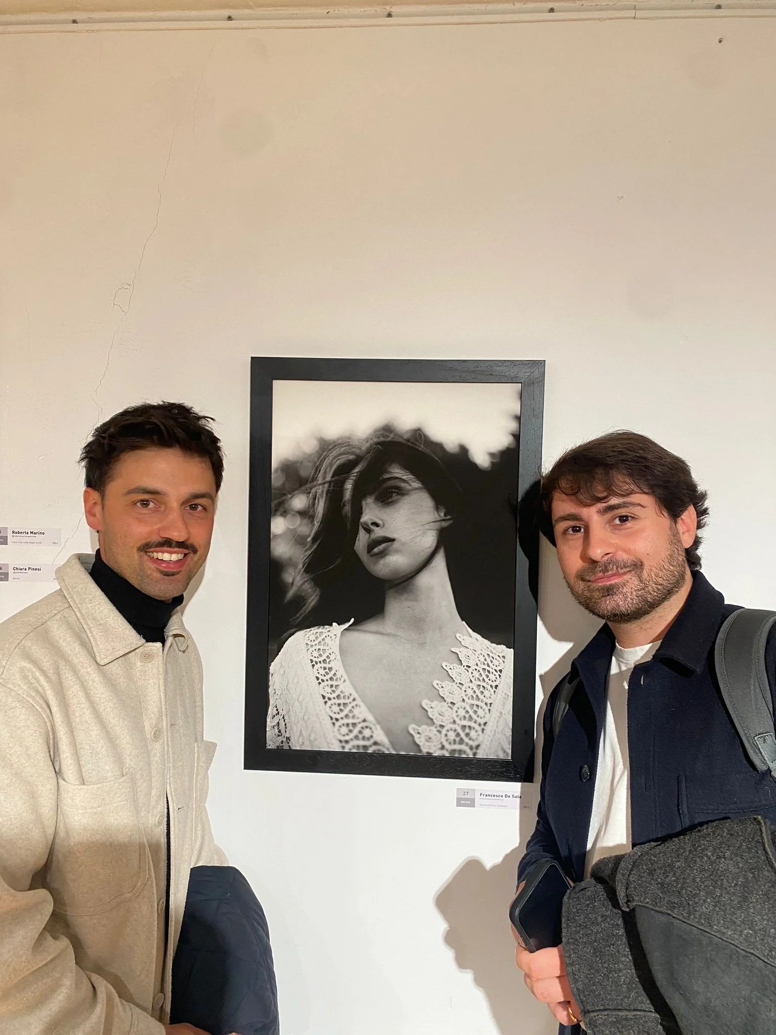 Due uomini sorridenti posano davanti a un quadro in bianco e nero di una donna con capelli mossi e abito ricamato, in una galleria d'arte.