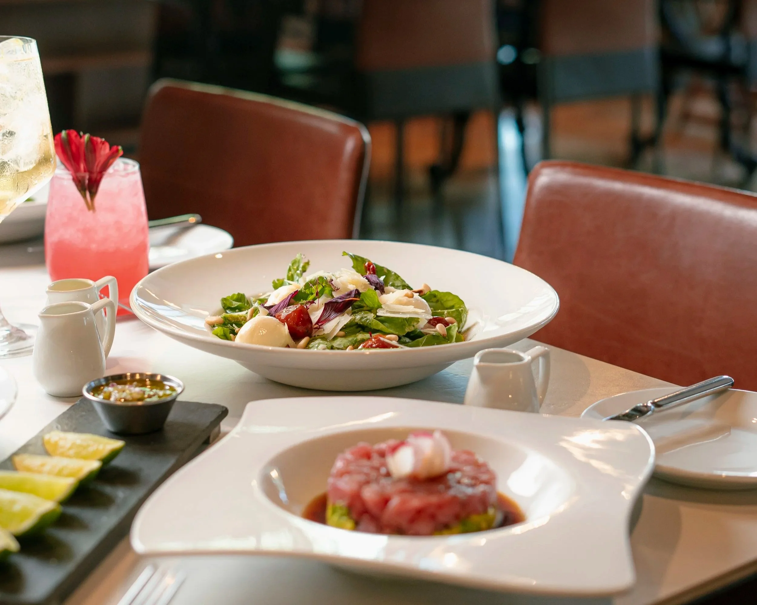 Una mesa con ensalada, rodajas de limón, salsas y comida en platos, en un restaurante con sillas de color marrón y ambiente informal.