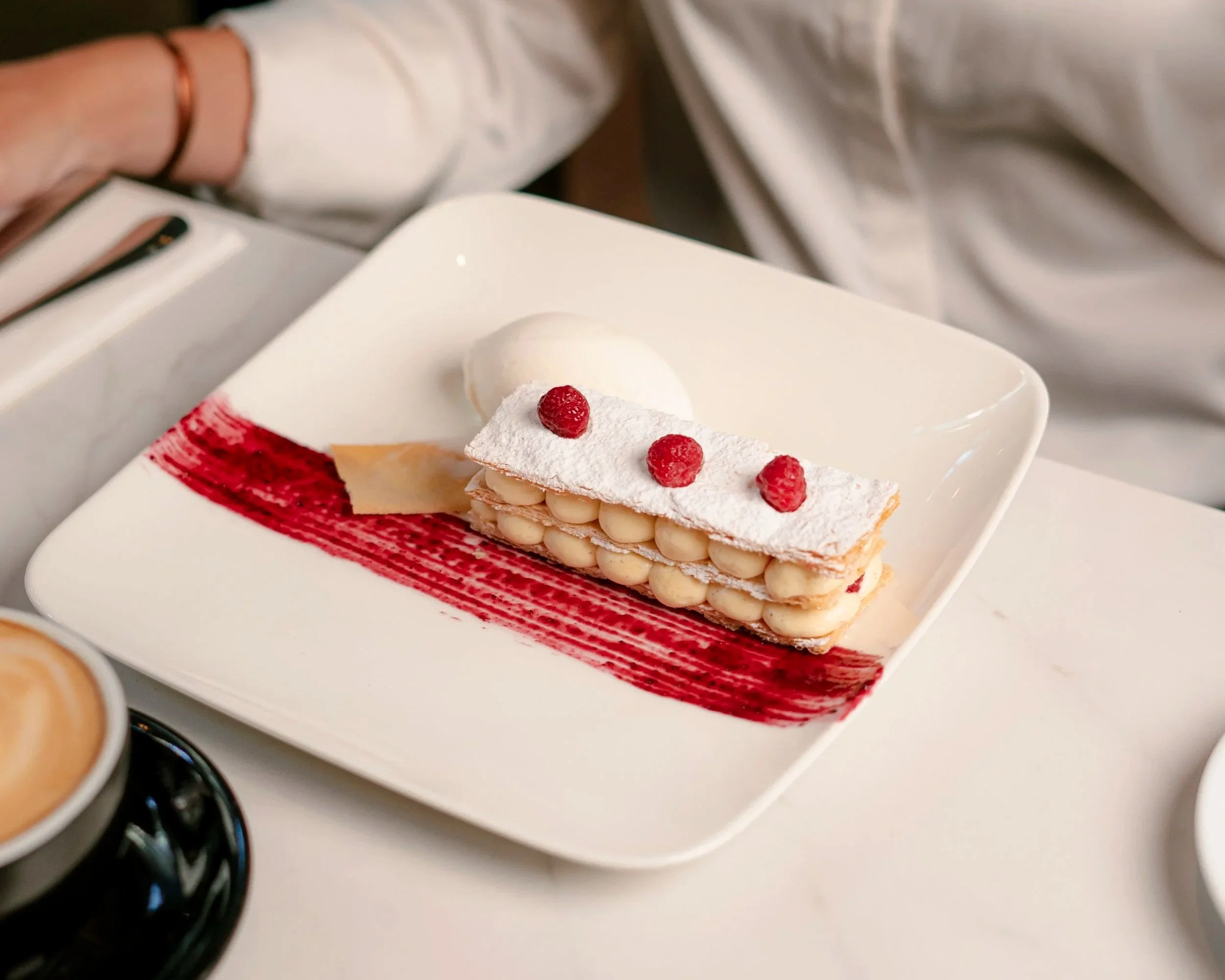 Postre de capas con crema, polvo de azúcar y frambuesas, decorado con salsa de frutos rojos en un plato blanco.