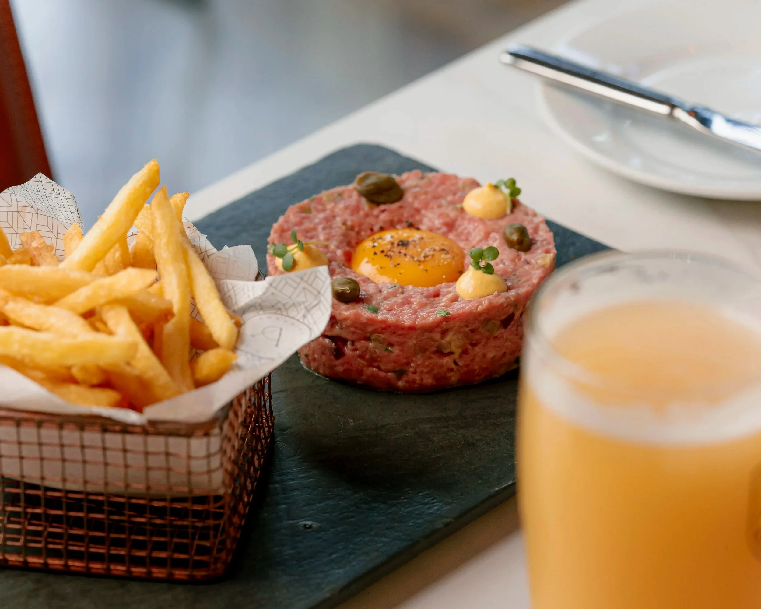Tartare de carne con huevo crudo, alcaparras y aderezo, acompañado de papas fritas en papel de aluminio, y en el fondo hay cubiertos y un vaso con bebida amarilla.