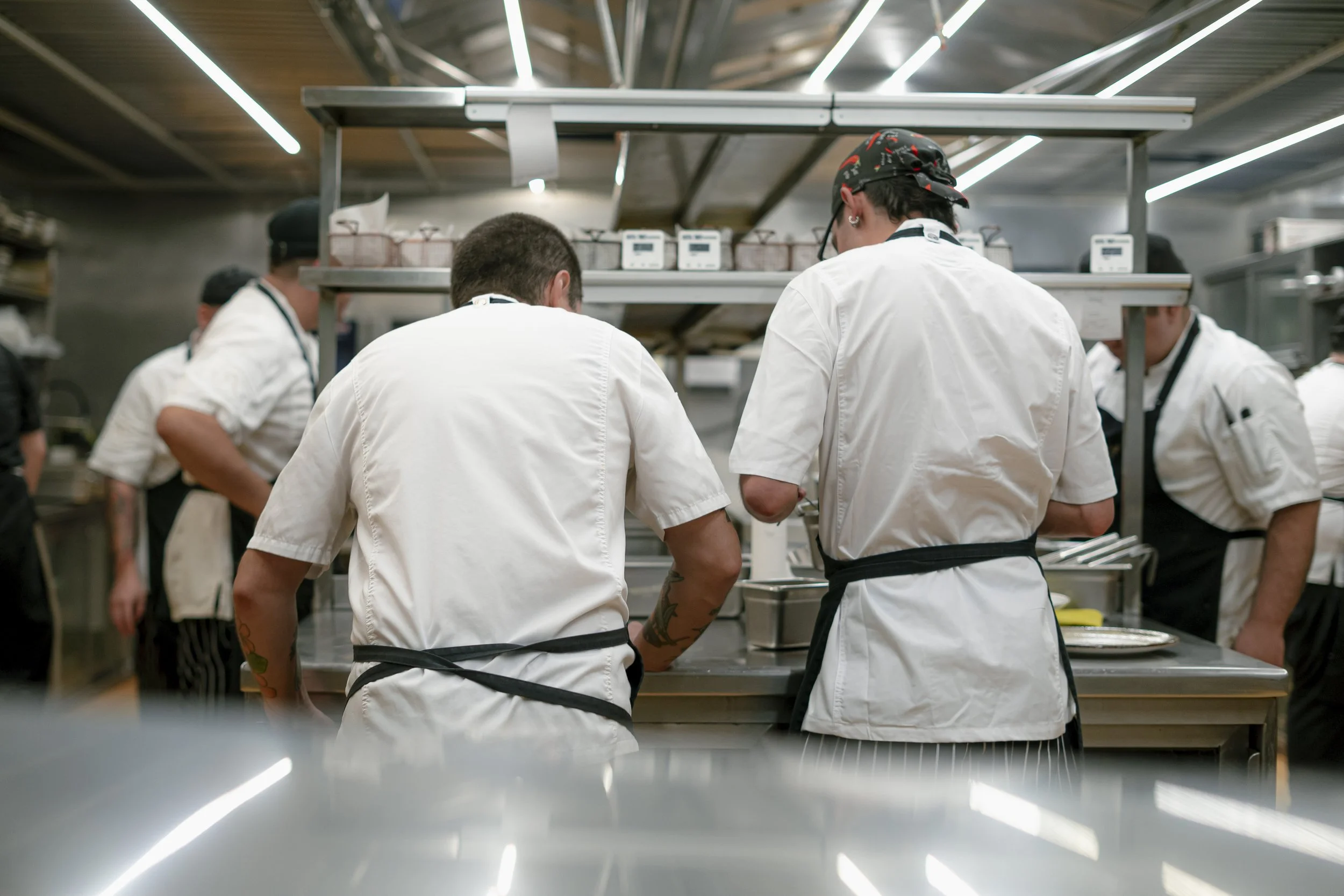 Grupo de chefs en la cocina, de espaldas, usando batas blancas y gorros, trabajando en la preparación de alimentos en una cocina industrial moderna.