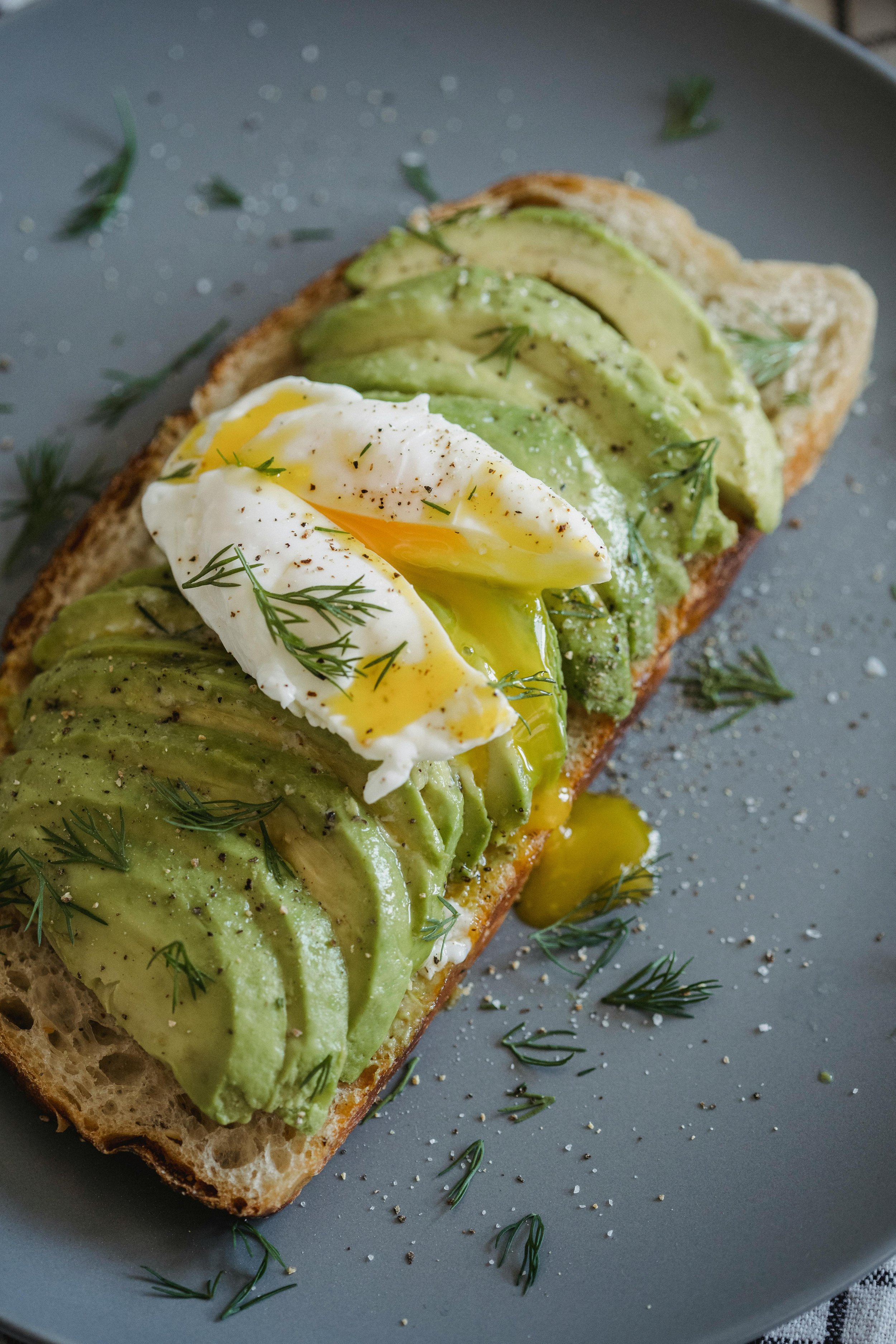 Tostada con aguacate en rebanadas, un huevo cocido parcialmente cortado, aceitunas, eneldo y pimienta en una superficie gris.