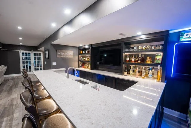 Home bar with a white countertop, barstools, a TV, and shelves with liquor bottles.
