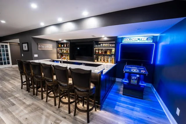 Home bar with a white countertop, black bar stools, illuminated shelves with liquor bottles, and an electronic basketball game with blue neon lighting.