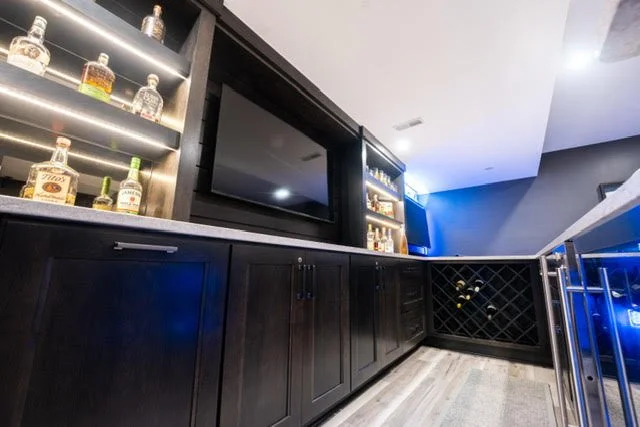 Home bar with black cabinetry, liquor bottles on illuminated shelves, a television, and a built-in wine rack.
