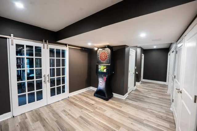Empty basement room with dark gray walls, light wood flooring, double French doors, a dartboard game, and white closet doors.
