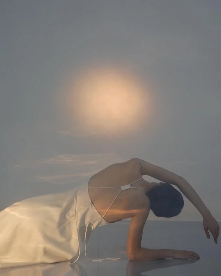 Een persoon in een witte jurk ligt op de grond met één arm gestrekt, met een reflectie of lichtvlek zichtbaar in de lucht erboven.