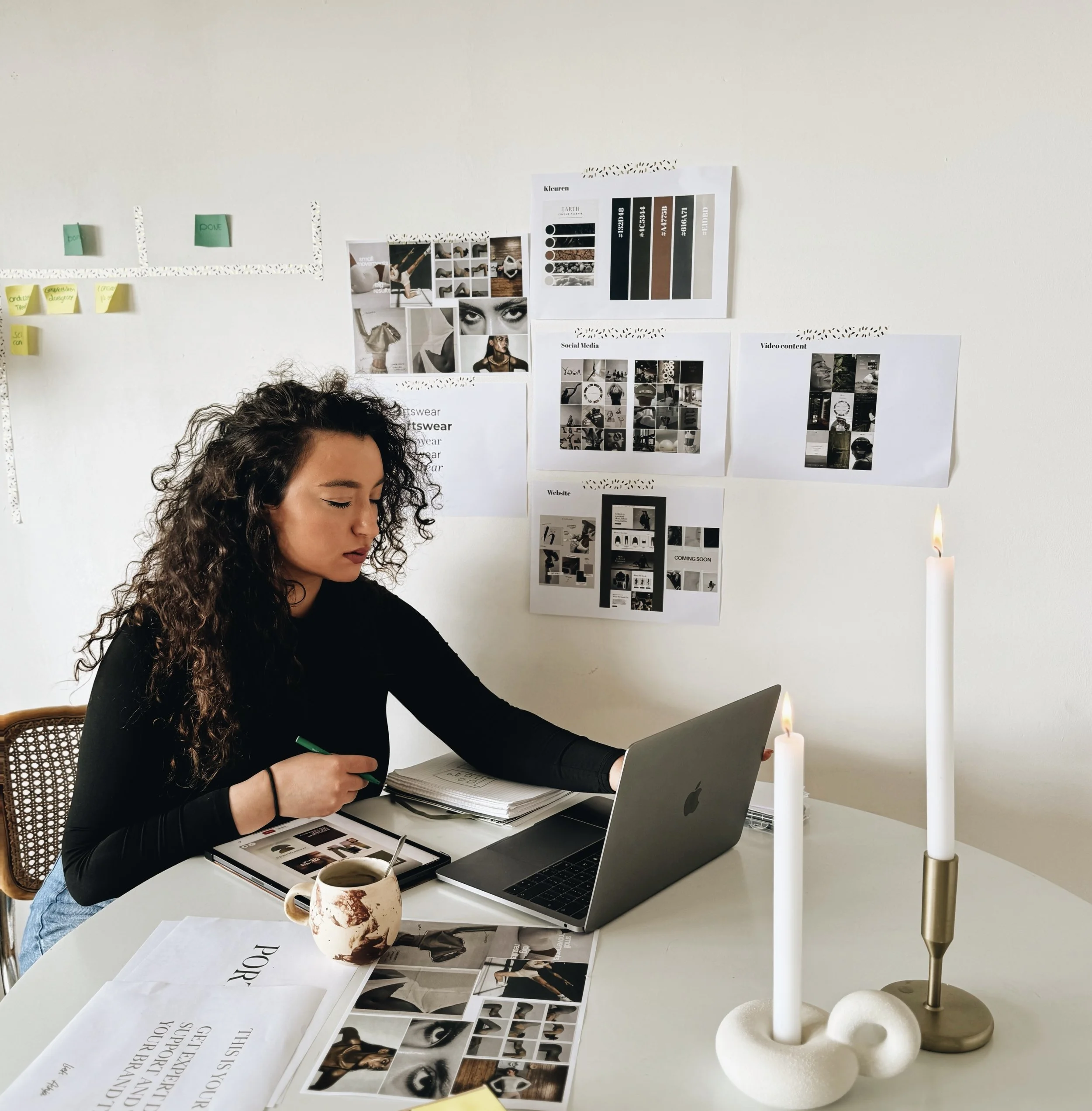 Vrouw werkt aan ontwerp of moodboard met laptop, notitieboekje, en foto's op tafel, achter haar post-its en moodboard met kleur en beeldcollages, in een lichte kamer.