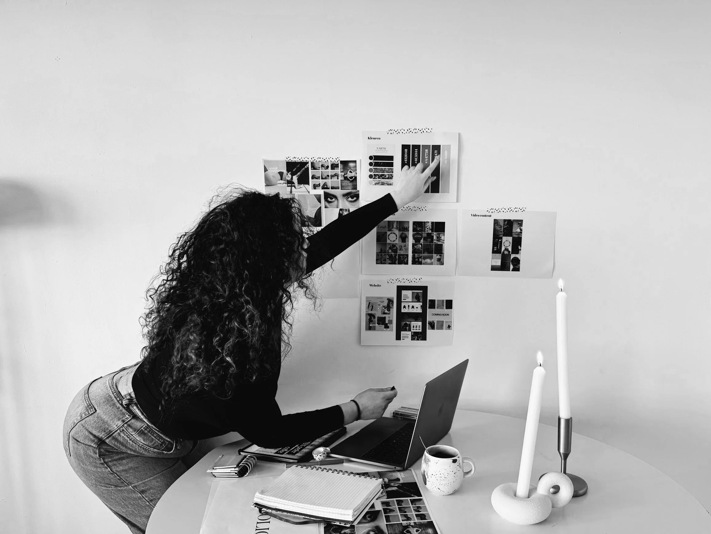 Een vrouw met krullend haar wijst op een prikbord met ontwerpen of moodboards, terwijl ze over een bureau met een open laptop, notitieboek, een kopje koffie, en decoratieve kaarsen buigt.