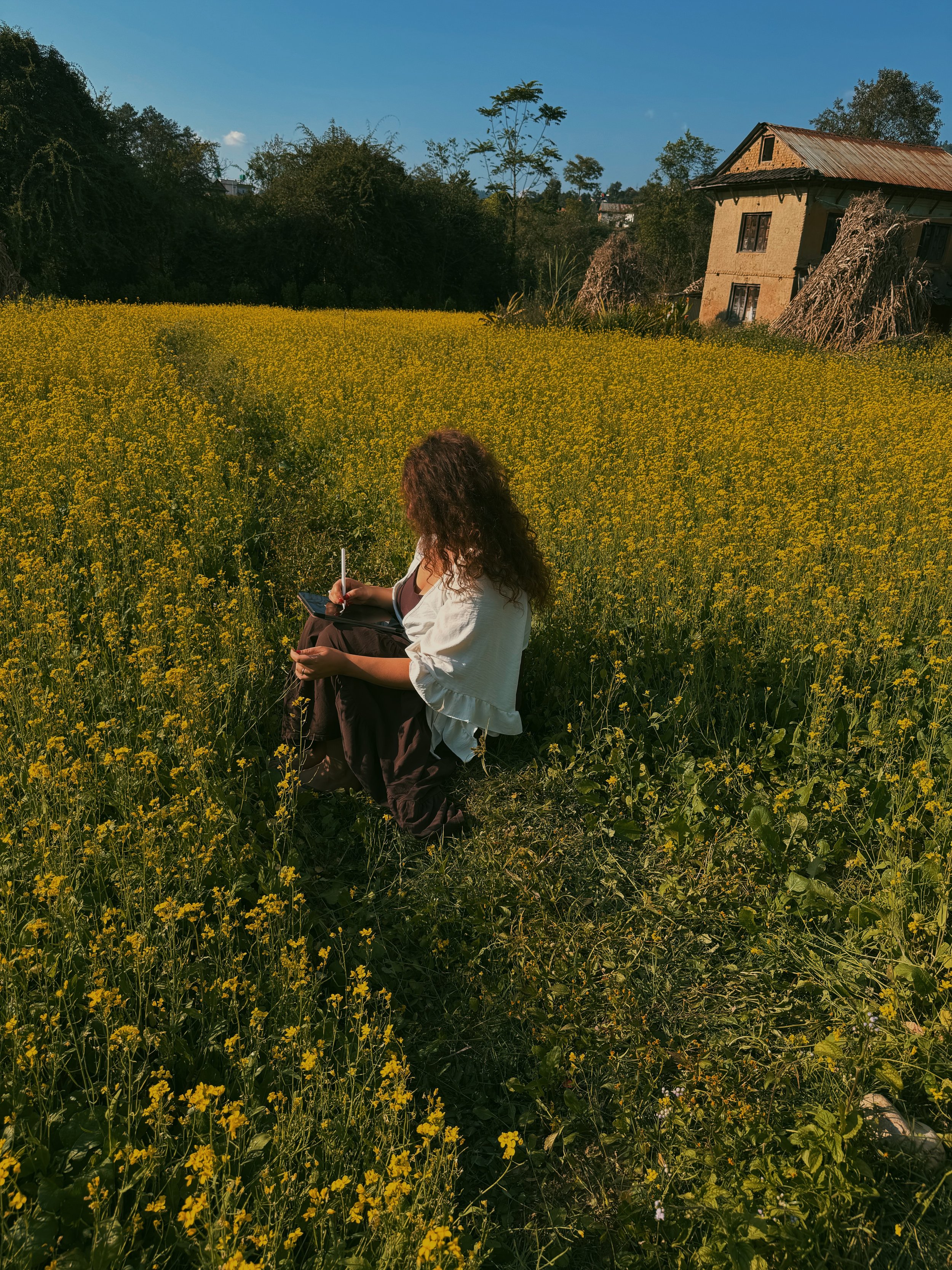 Vrouw zit in een veld met gele bloemen, schrijft in een notitieboekje, met een huis en bomen op de achtergrond onder een blauwe hemel.