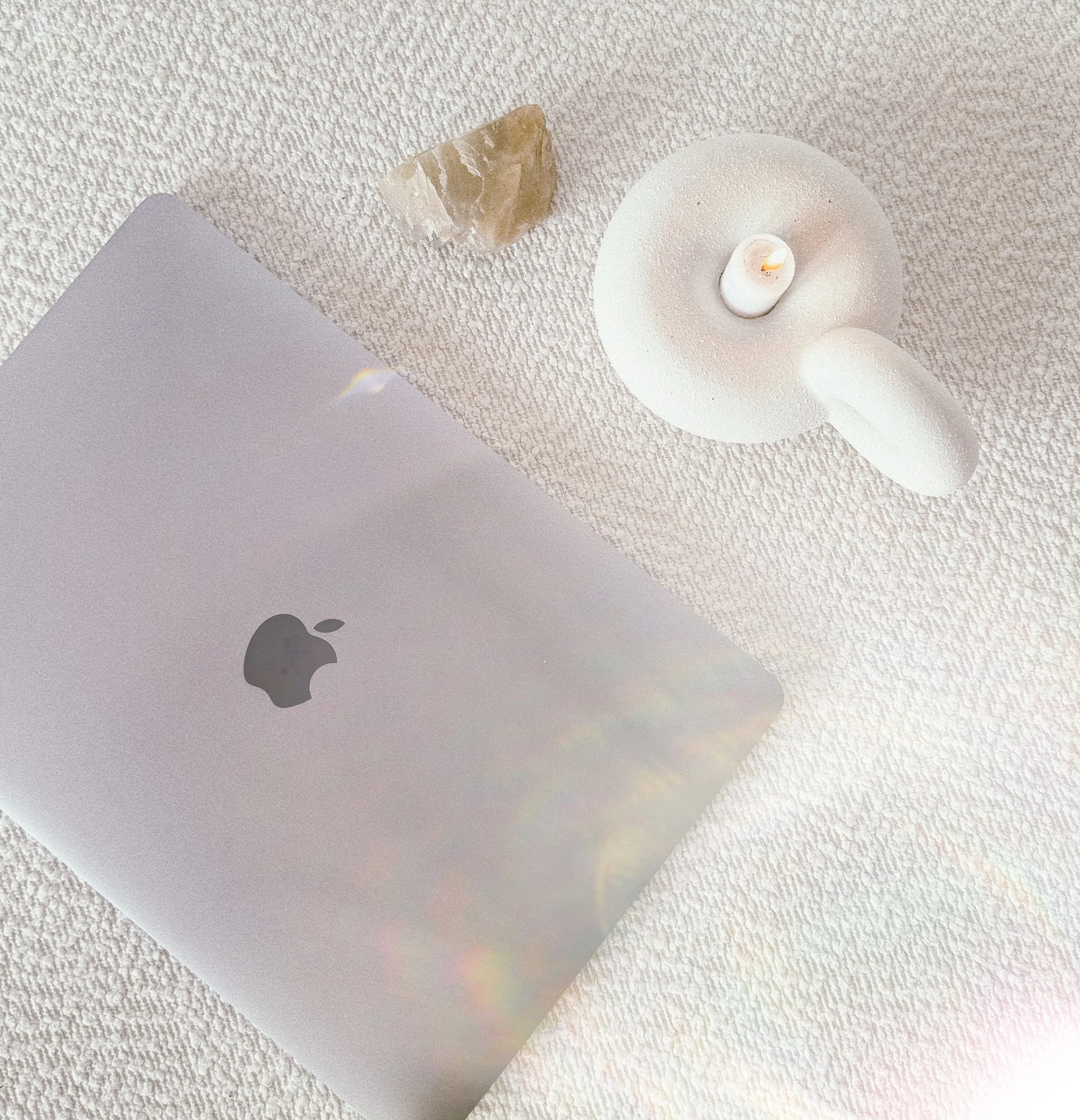 Een closed silver MacBook op een zachte, lichtgekleurde stoffen ondergrond, naast een handgemaakte witte kaars en een stuk rozekwarts in een decoratieve setting.