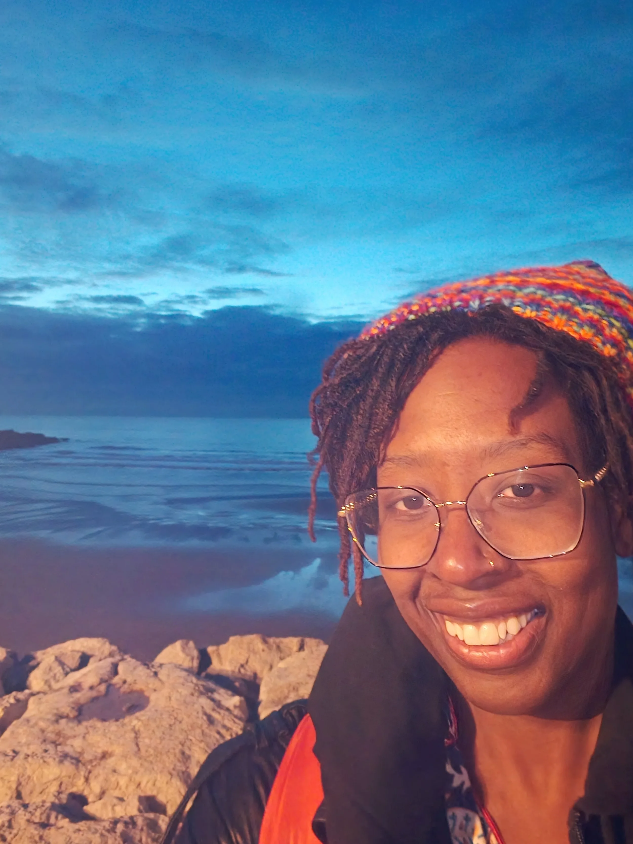 A woman with glasses and colorful braided hair smiling at the camera, standing outdoors near a beach with rocks, ocean, and a cloudy sky at sunset in the background.