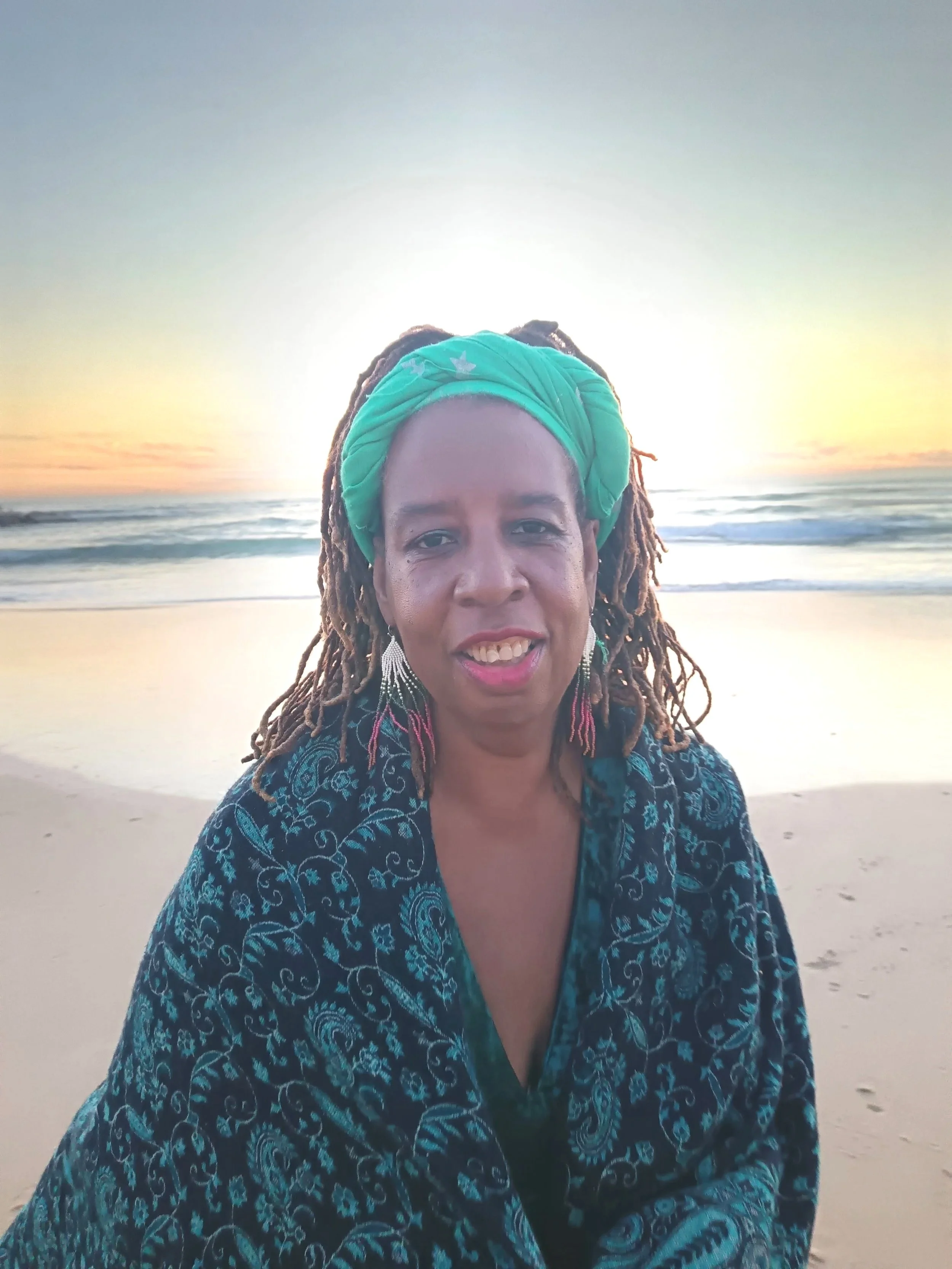 woman with a green headwrap and earrings, sitting on the beach at sunset