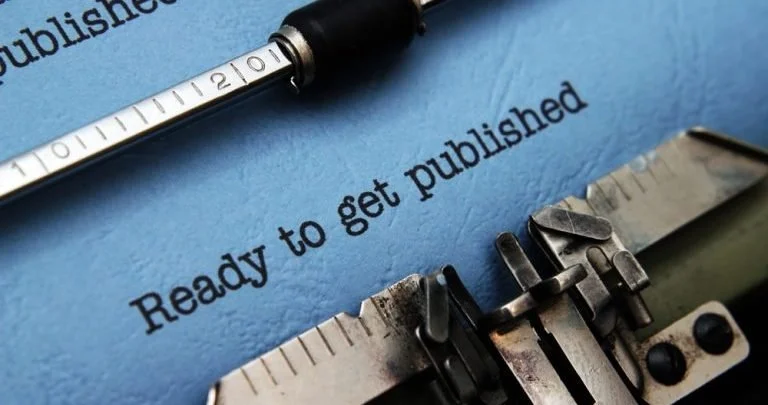 Close-up of a typewriter with the phrase 'Ready to get published' typed on blue paper, accompanied by a white and black pen.