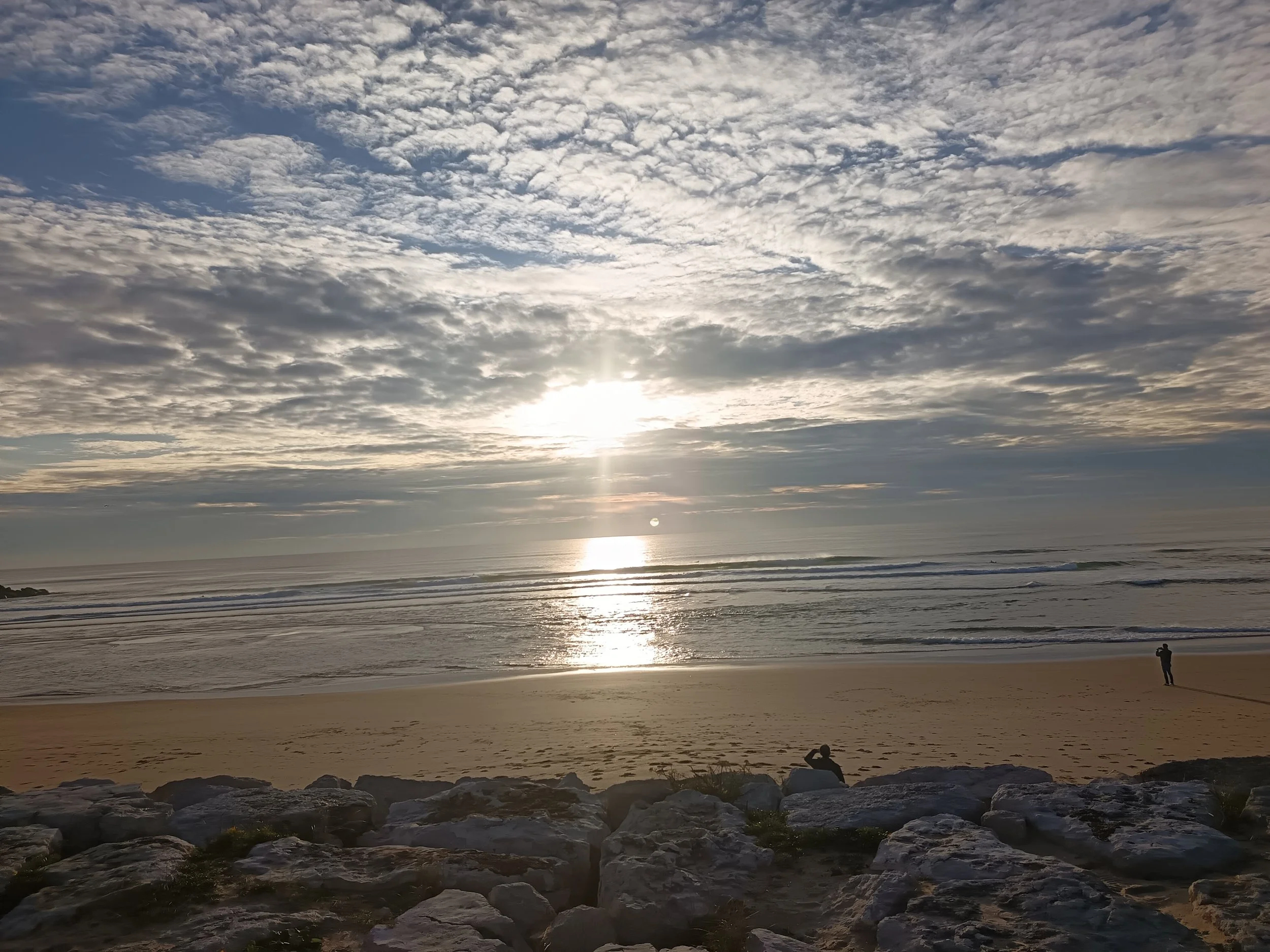Sunset over the ocean with scattered clouds, sandy beach, rocks in the foreground, and two people on the beach, one standing and one sitting.