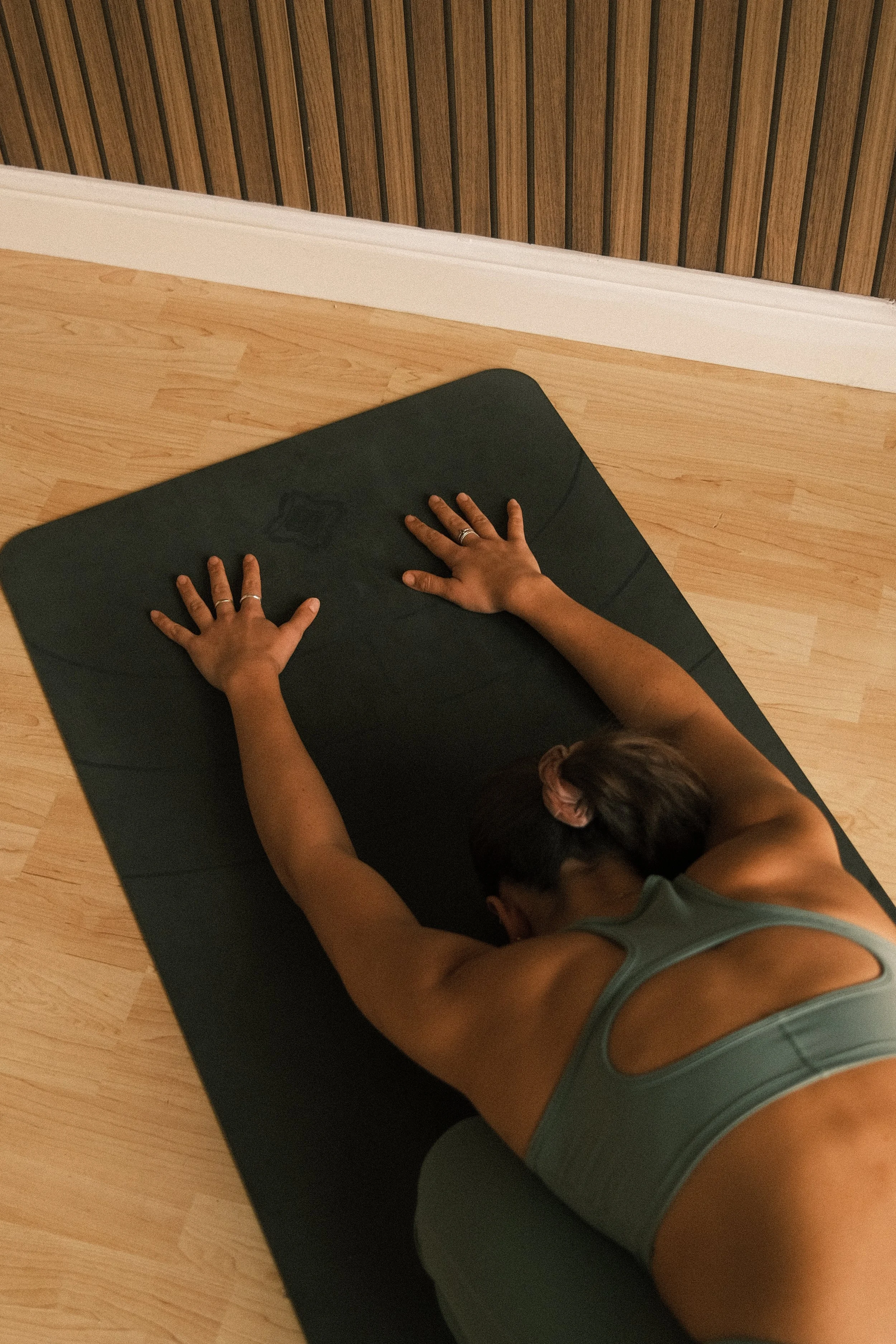 Person practicing yoga or stretching on a black mat indoors, with wood flooring and vertical wood paneling on the wall.