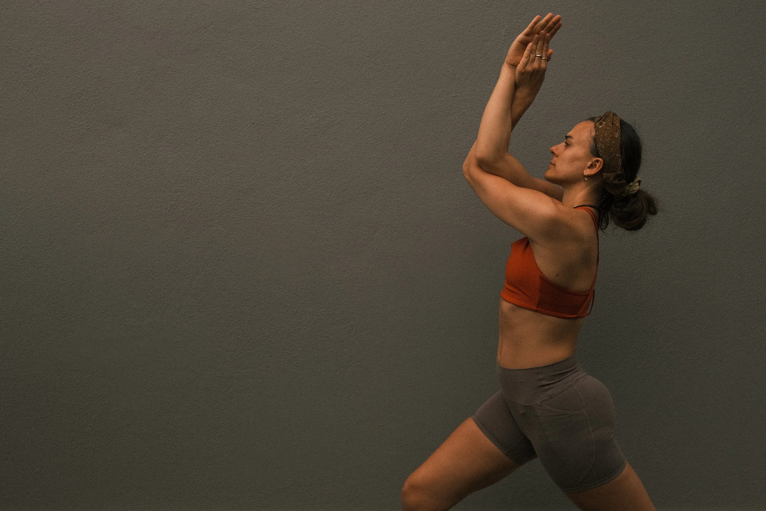 A woman practicing yoga or meditation against a plain grey wall, wearing a brown headband, an orange sports bra, and grey shorts, in a side profile pose with hands pressed together above her head.