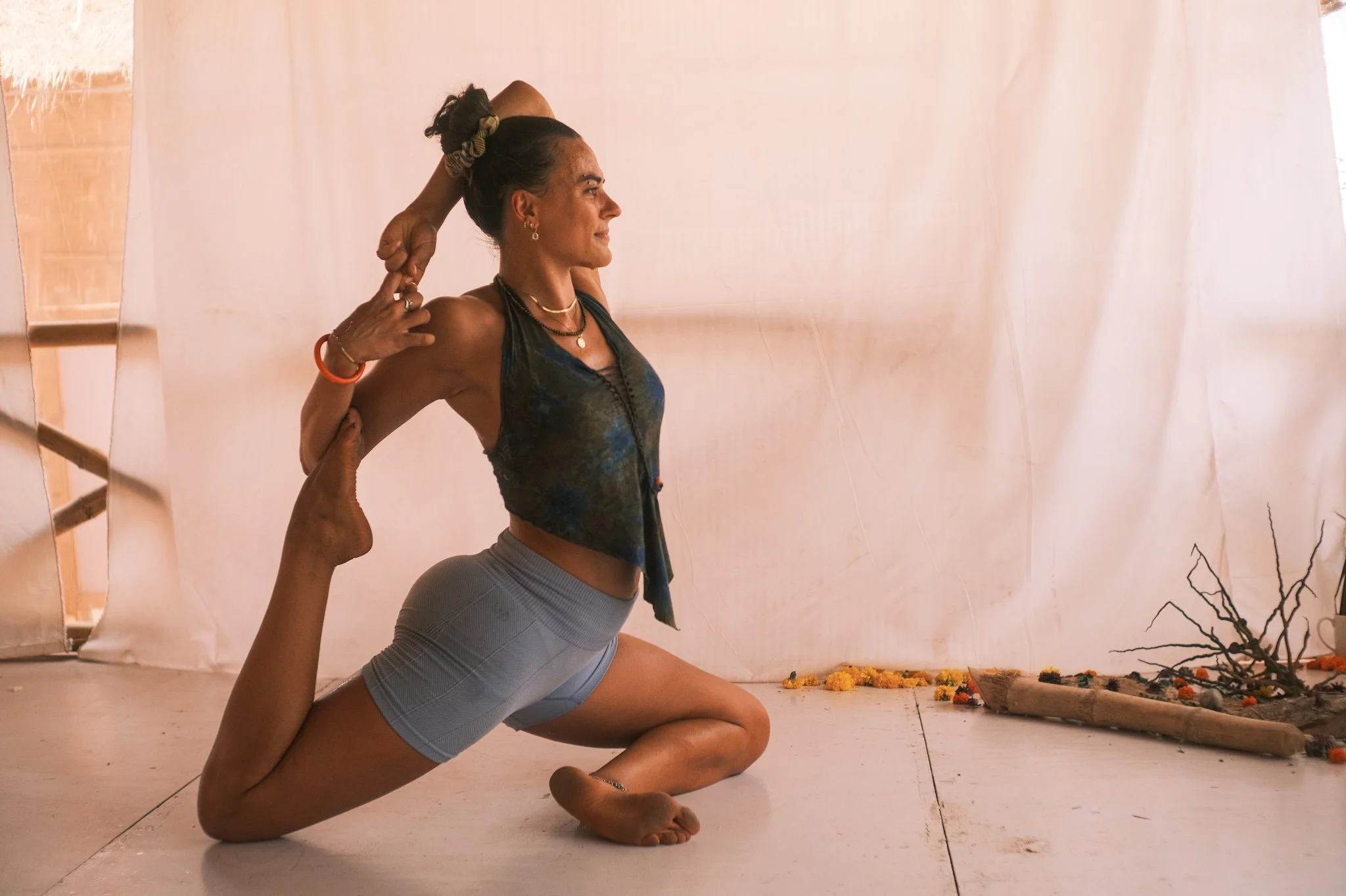 A woman practicing yoga in a king pigeon pose, stretching her arm behind her head and holding her foot with her hand, indoors with a light-colored curtain background.